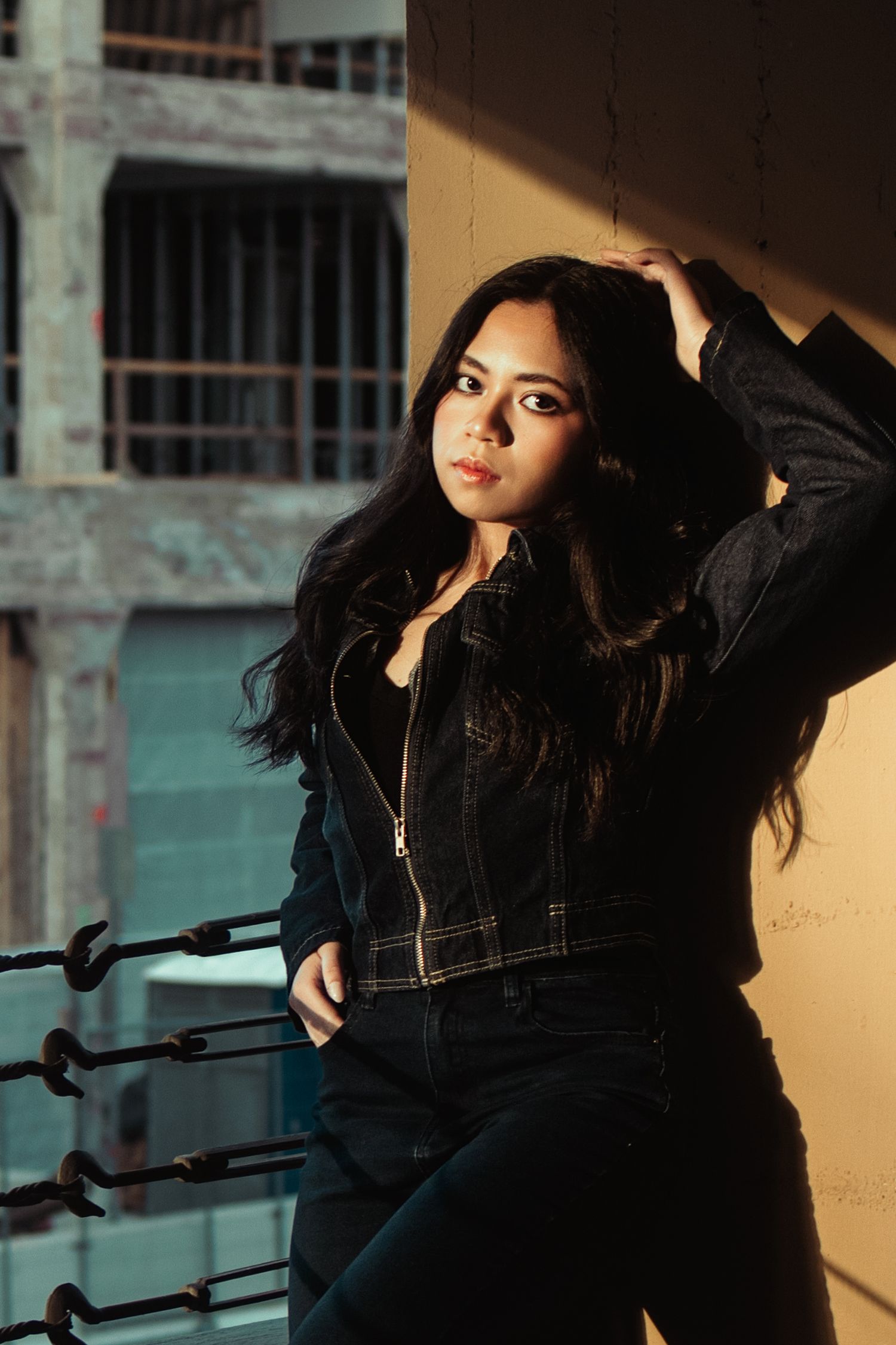 Young woman in black denim jacket poses confidently on urban rooftop balcony with long dark hair in sunlight.