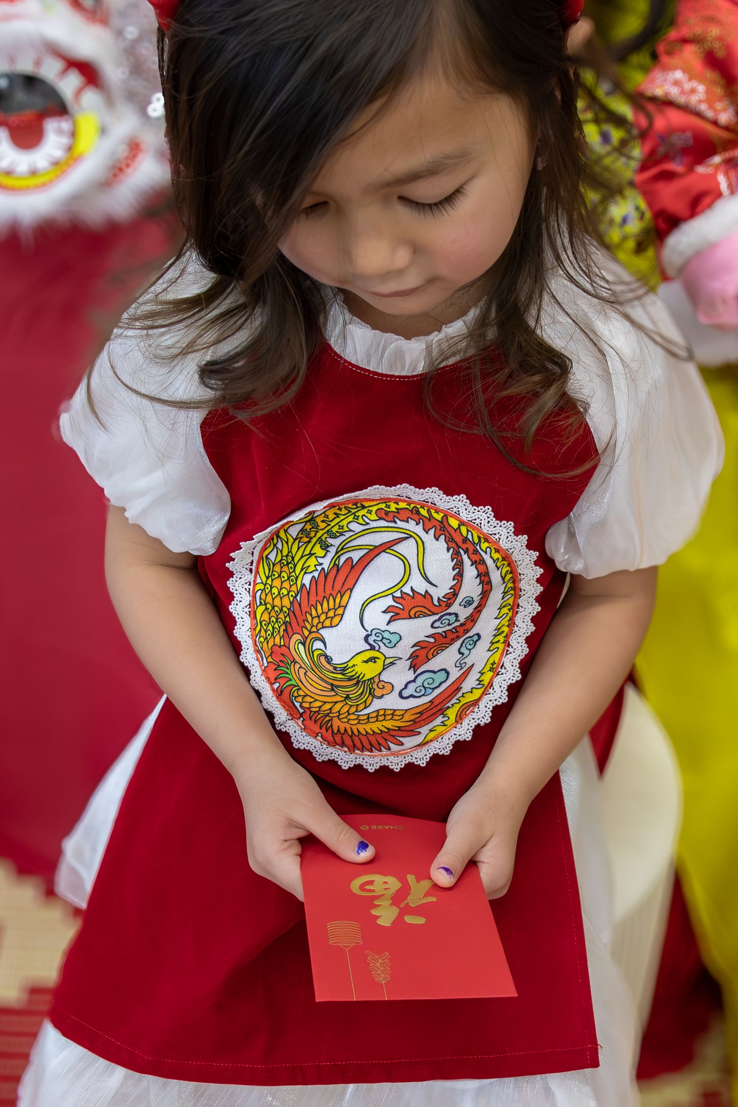 Young girl in red and white Chinese dress holding a red envelope during Lunar New Year celebration.