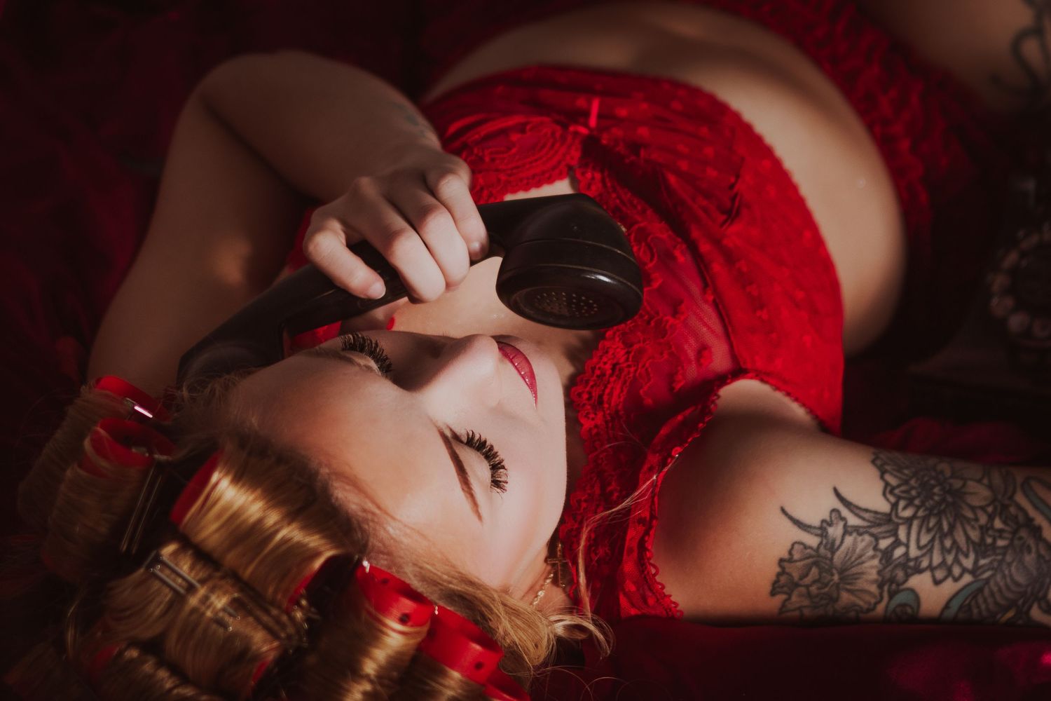 Woman in red lace lingerie lying down, wearing dark goggles and gold bracelets, with floral tattoo on arm.