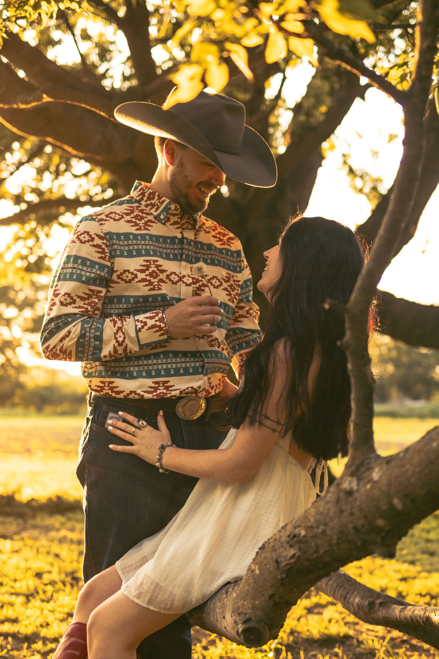 Couple embracing near a tree at golden sunset, man in cowboy hat and Aztec print shirt with woman in white dress.