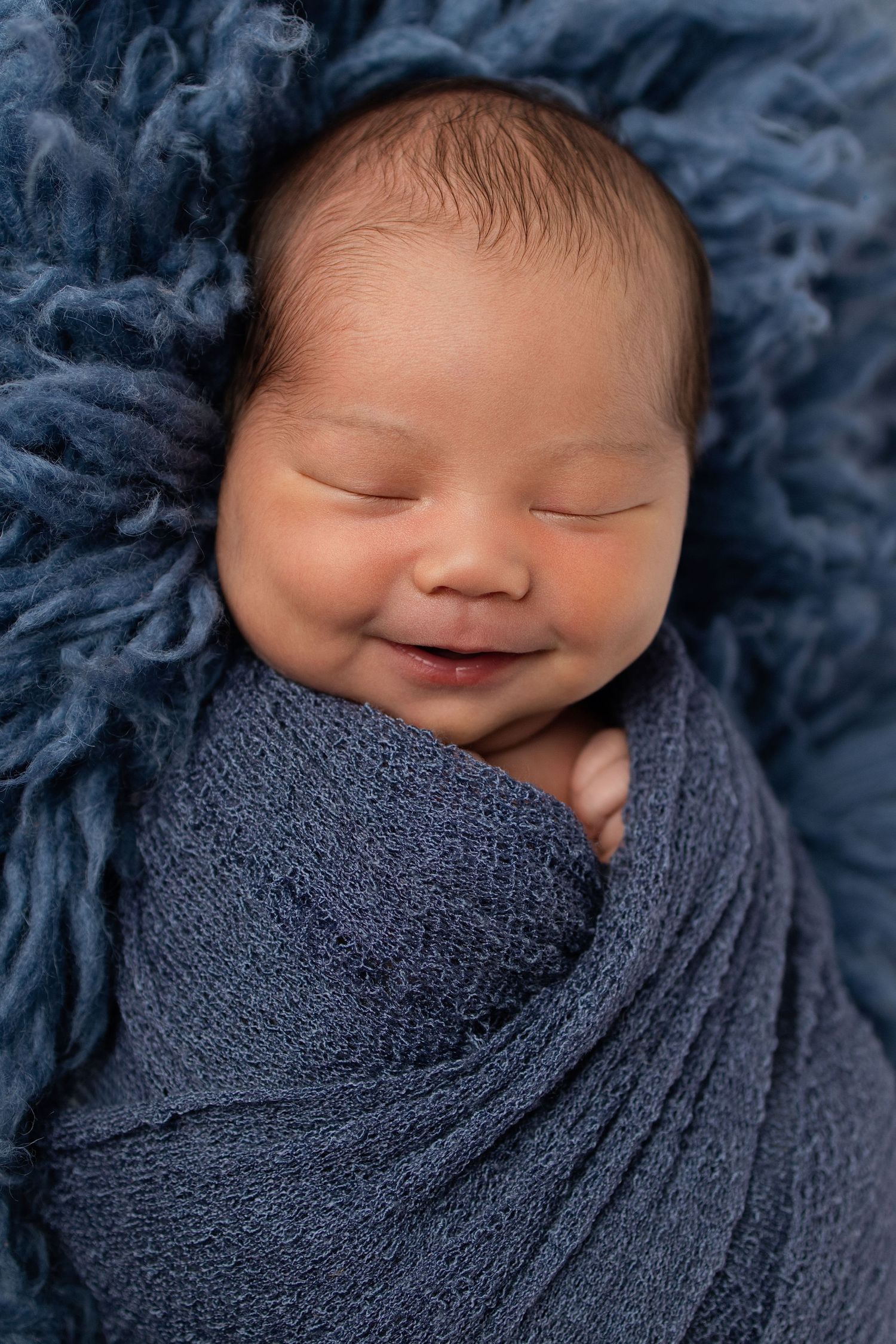 Smiling newborn baby swaddled in a blue wrap, peacefully sleeping on a soft blue fluffy blanket.