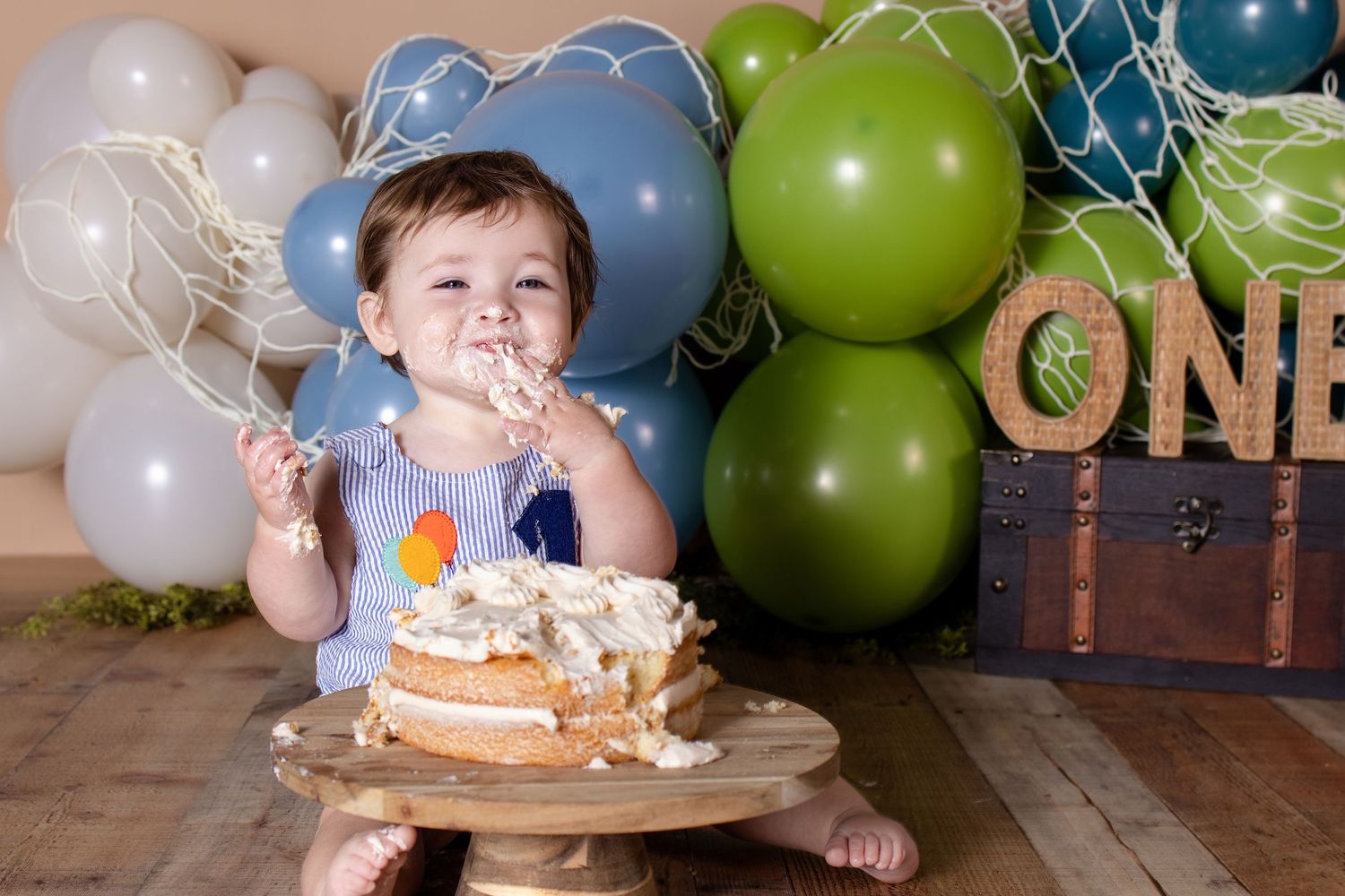 Adorable baby in striped romper eating smash cake at first birthday party with blue and green balloon decorations.