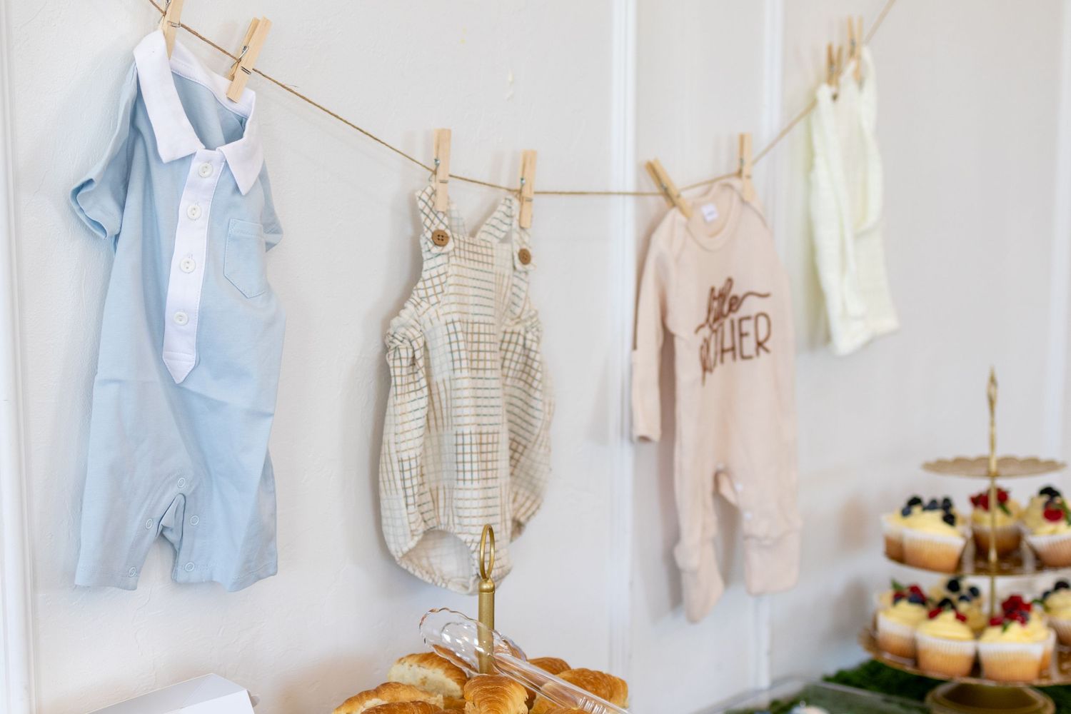 Baby clothes hanging on a clothesline at a gender neutral baby shower with cupcakes and pastries below.