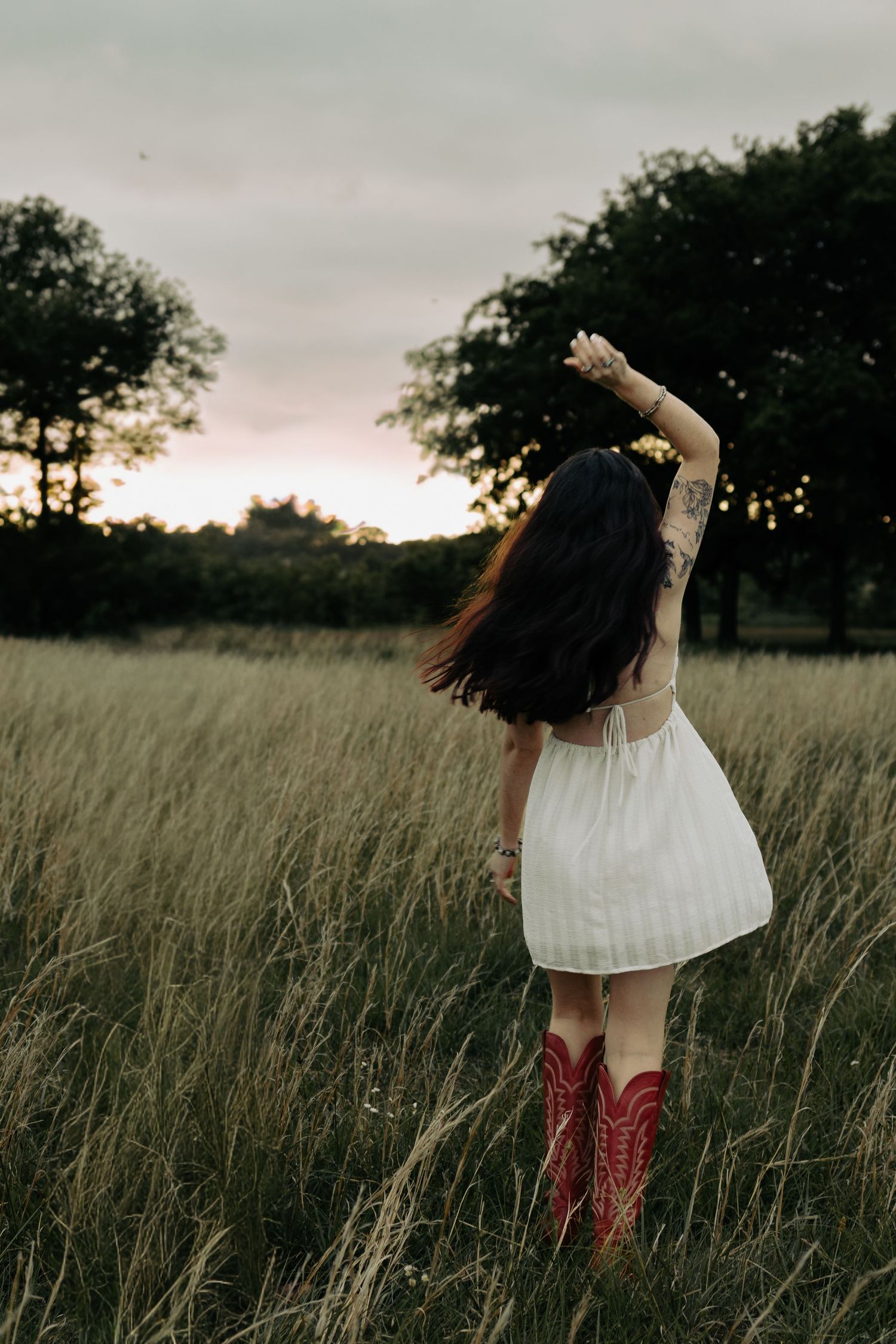 Woman in white dress and red cowboy boots standing in tall grass field at sunset with arms raised.
