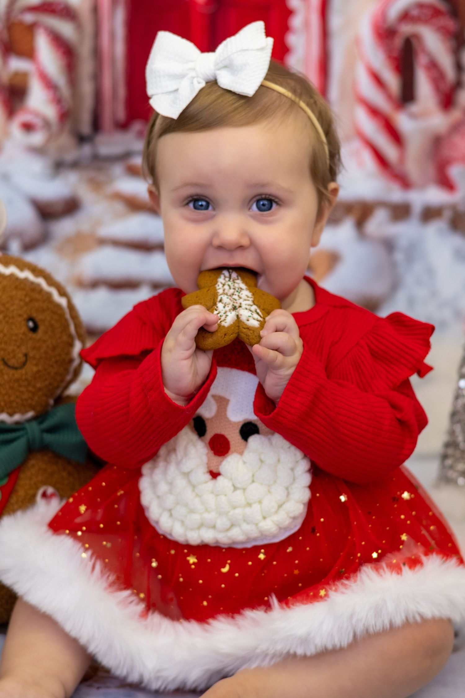 Adorable baby girl in red Santa dress eating a gingerbread cookie with white bow headband at Christmas.