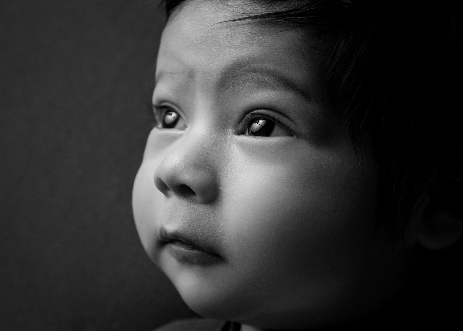 Black and white portrait of a curious baby looking up with wide expressive eyes against a dark background.
