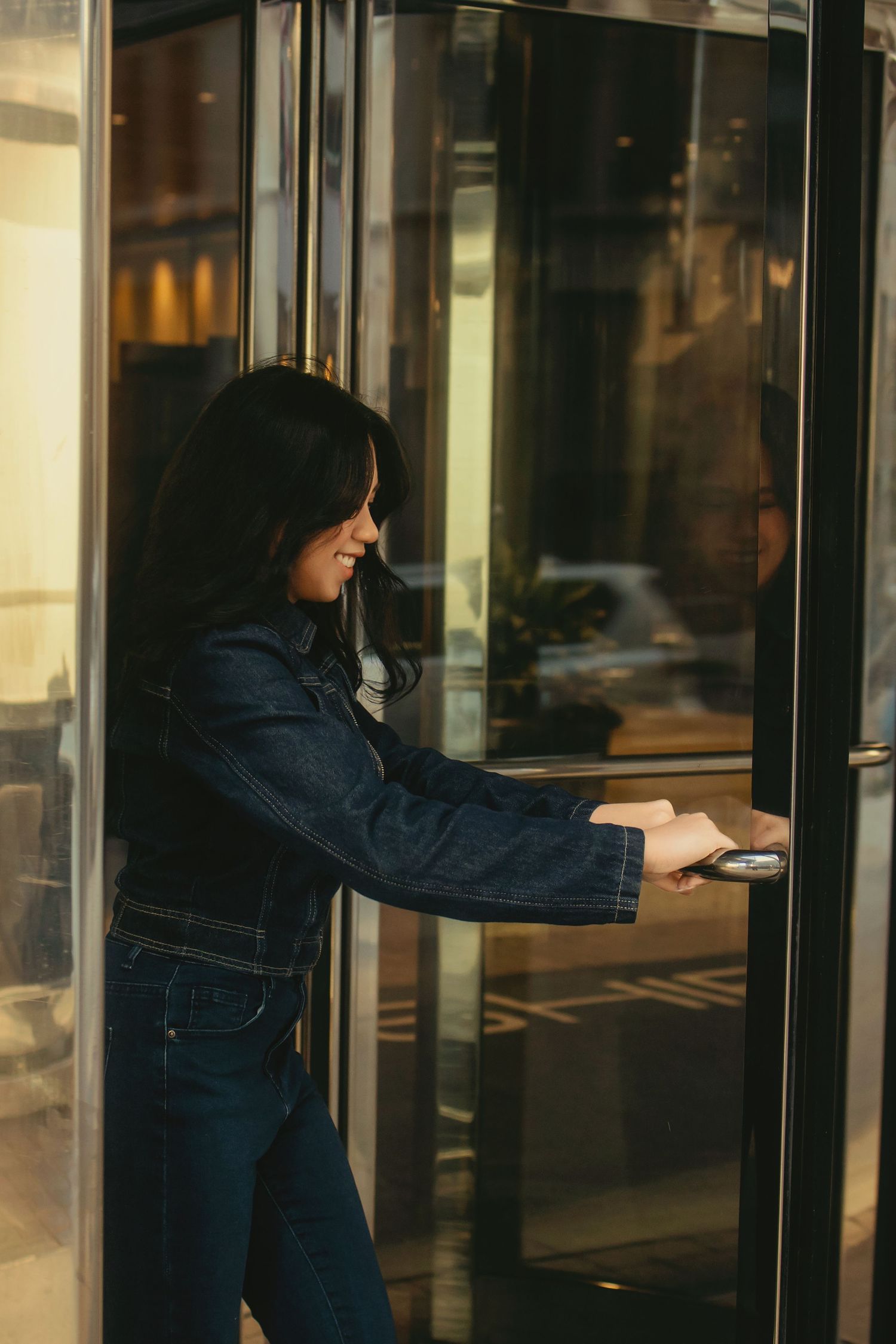 Smiling woman in denim jacket opens glass revolving door bathed in warm golden sunlight.