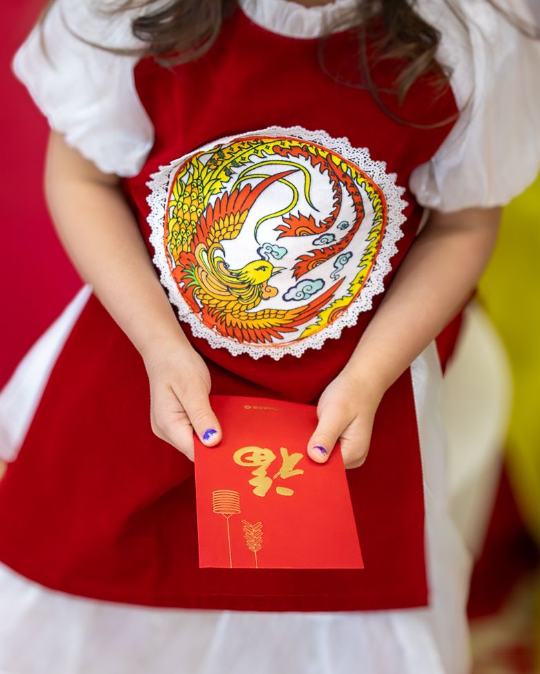 Girl in red dress holds decorative phoenix plate and red envelope for Chinese New Year celebration.