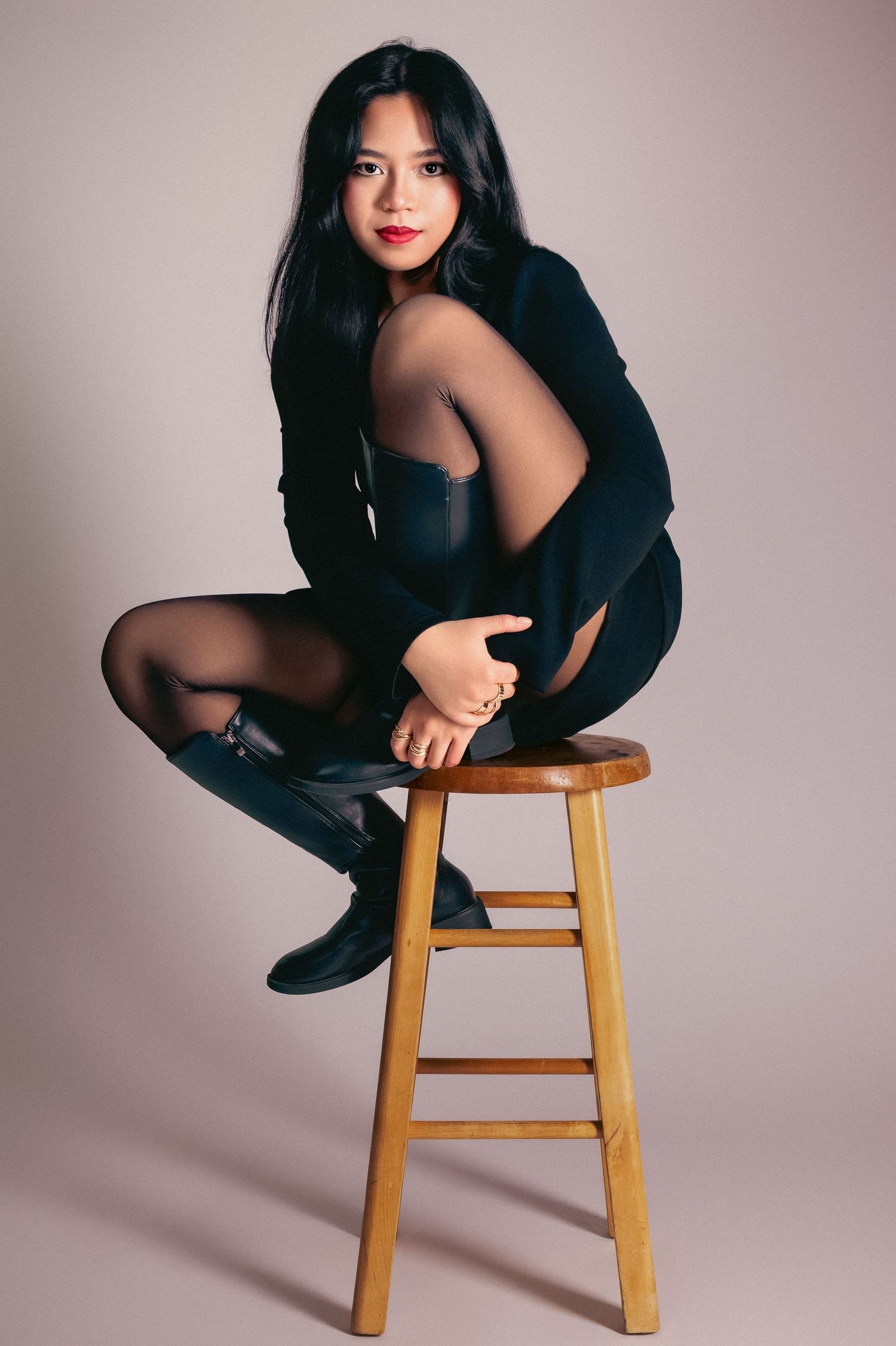 Young Asian woman in black outfit and boots poses on wooden stool against neutral background.