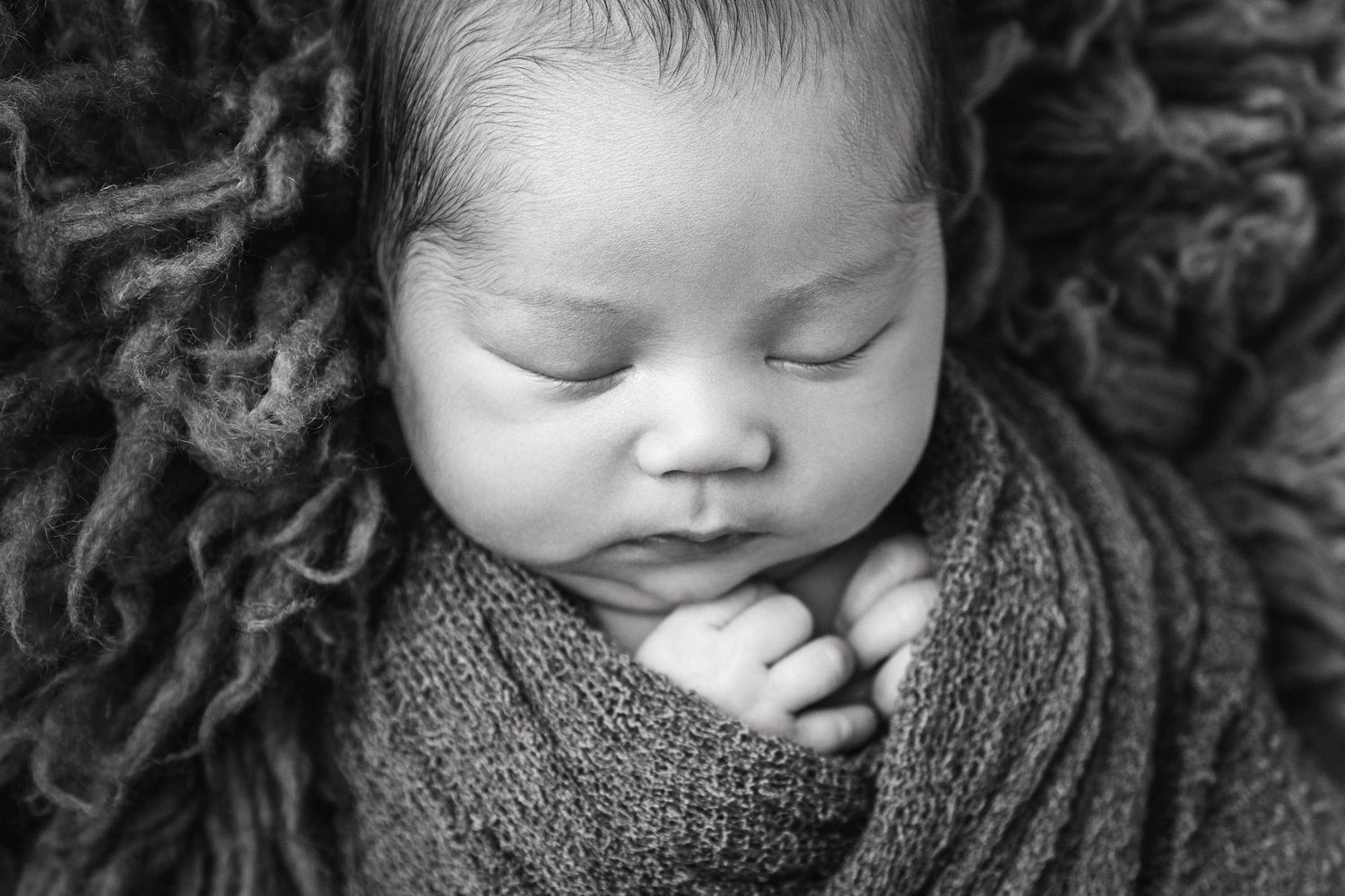 Black and white newborn baby sleeping peacefully, swaddled in soft knit wrap with tiny hands visible.