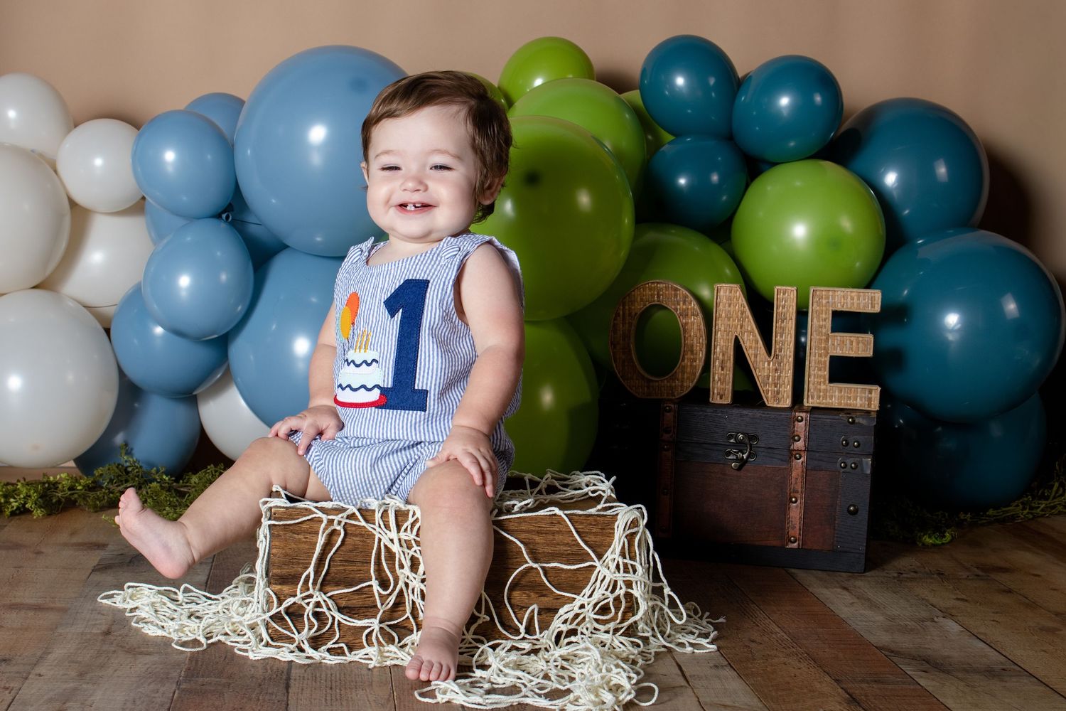 Smiling baby boy in blue romper sits on wooden crate with ONE sign and green and blue balloon garland backdrop.