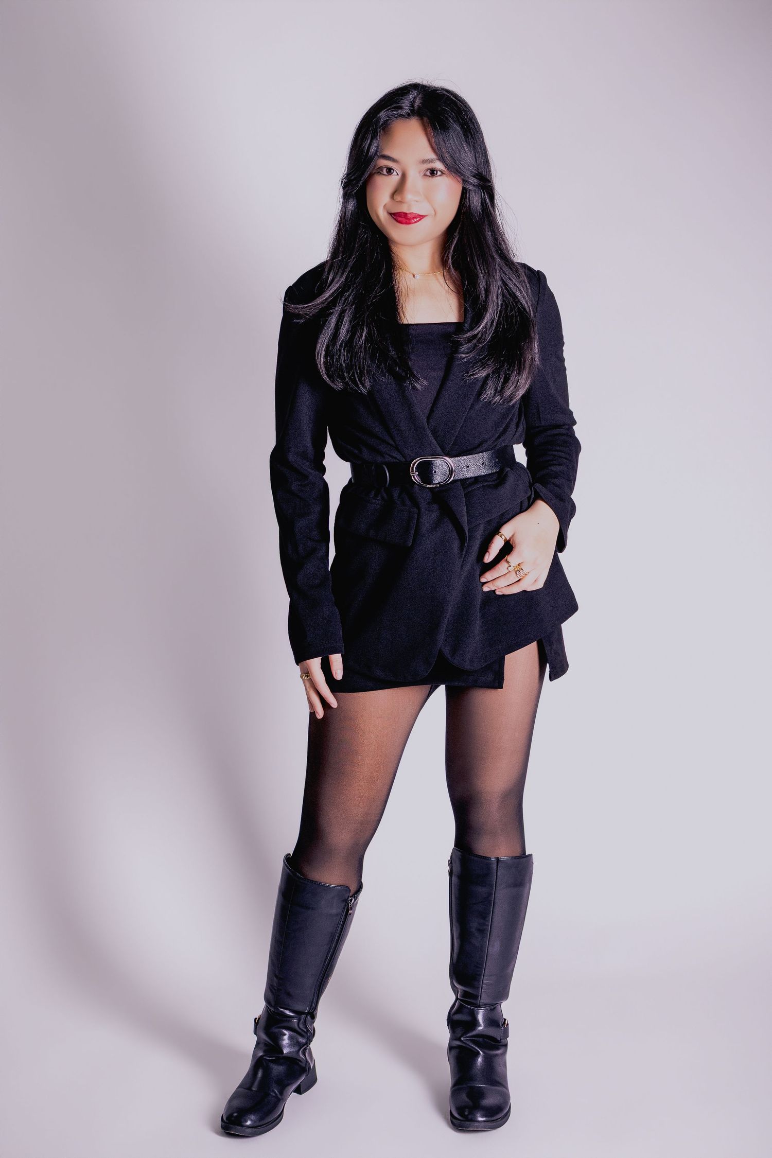 Young Asian woman in black belted blazer dress, tights, and knee-high boots posing confidently in studio.