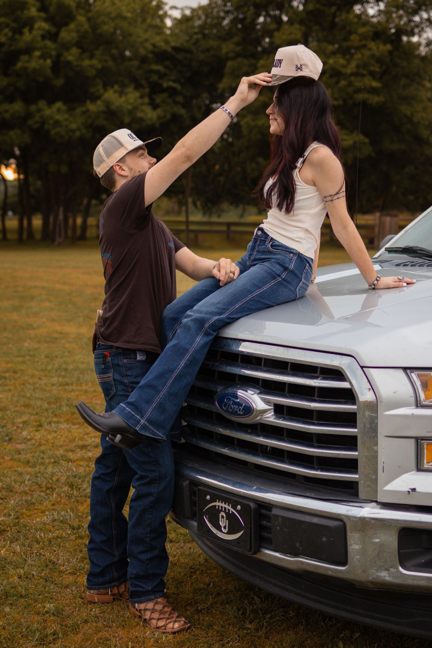 Couple sharing a playful moment on a Ford truck hood at dusk, woman sitting on hood in flare jeans.