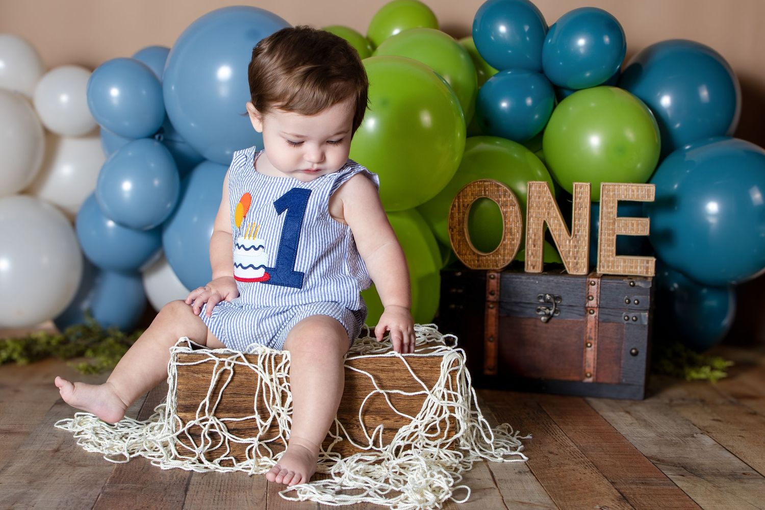 Baby boy in blue romper with number one sits by ONE sign surrounded by blue and green balloons for first birthday.