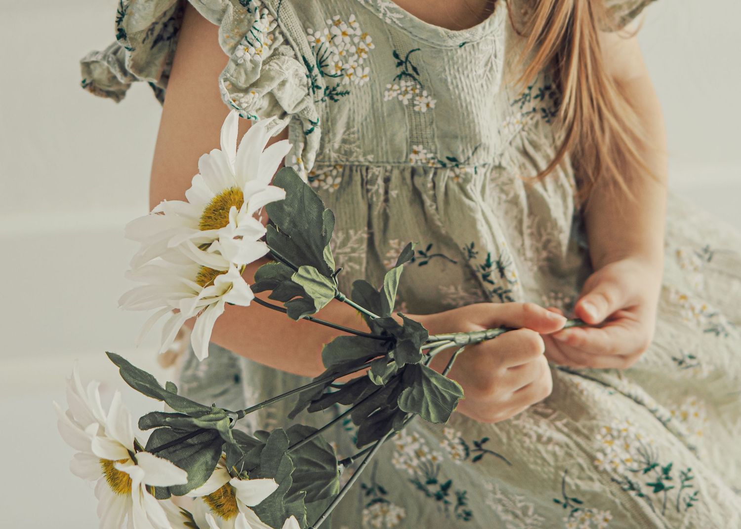 Young girl in floral dress holding white daisy flowers, soft light background.