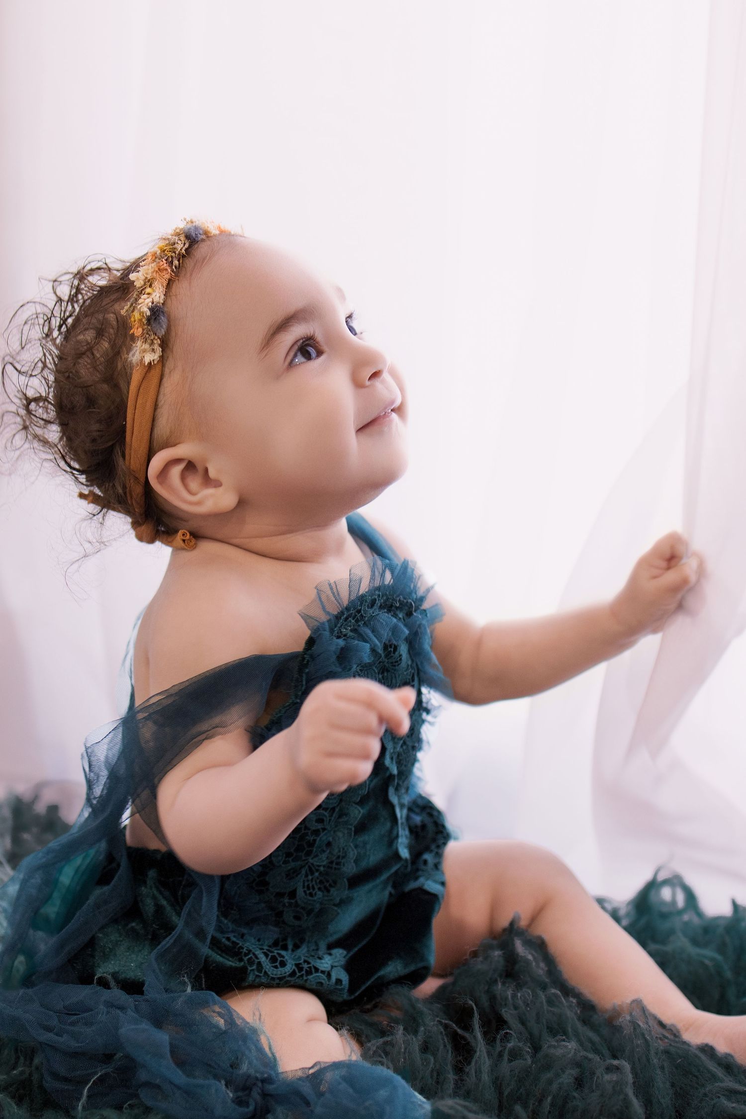 Adorable baby girl in teal tulle dress and gold floral headband sitting by white curtain looking up.