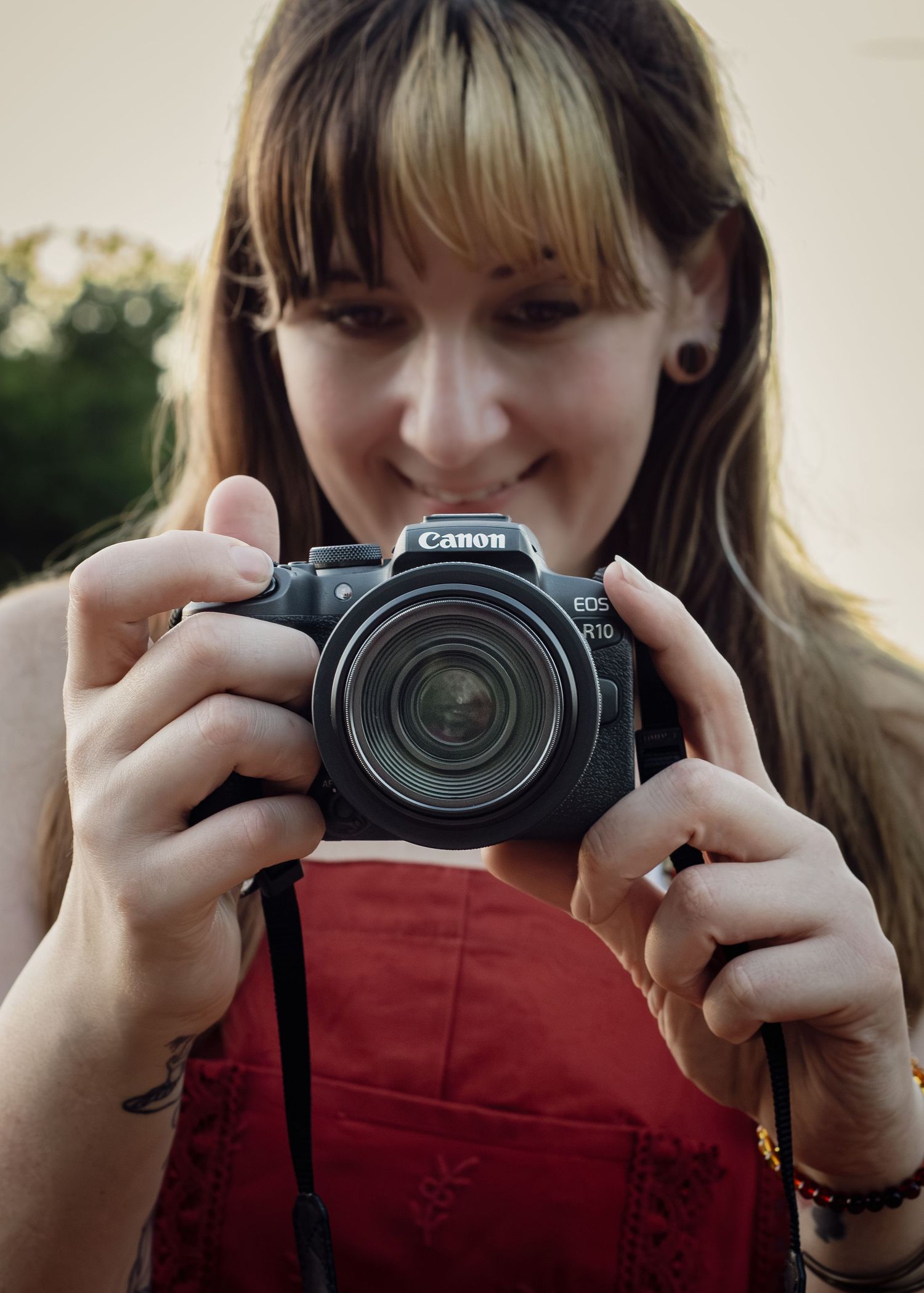 A smiling young woman with bangs holds a Canon EOS R10 camera up to photograph, wearing a red top outdoors.
