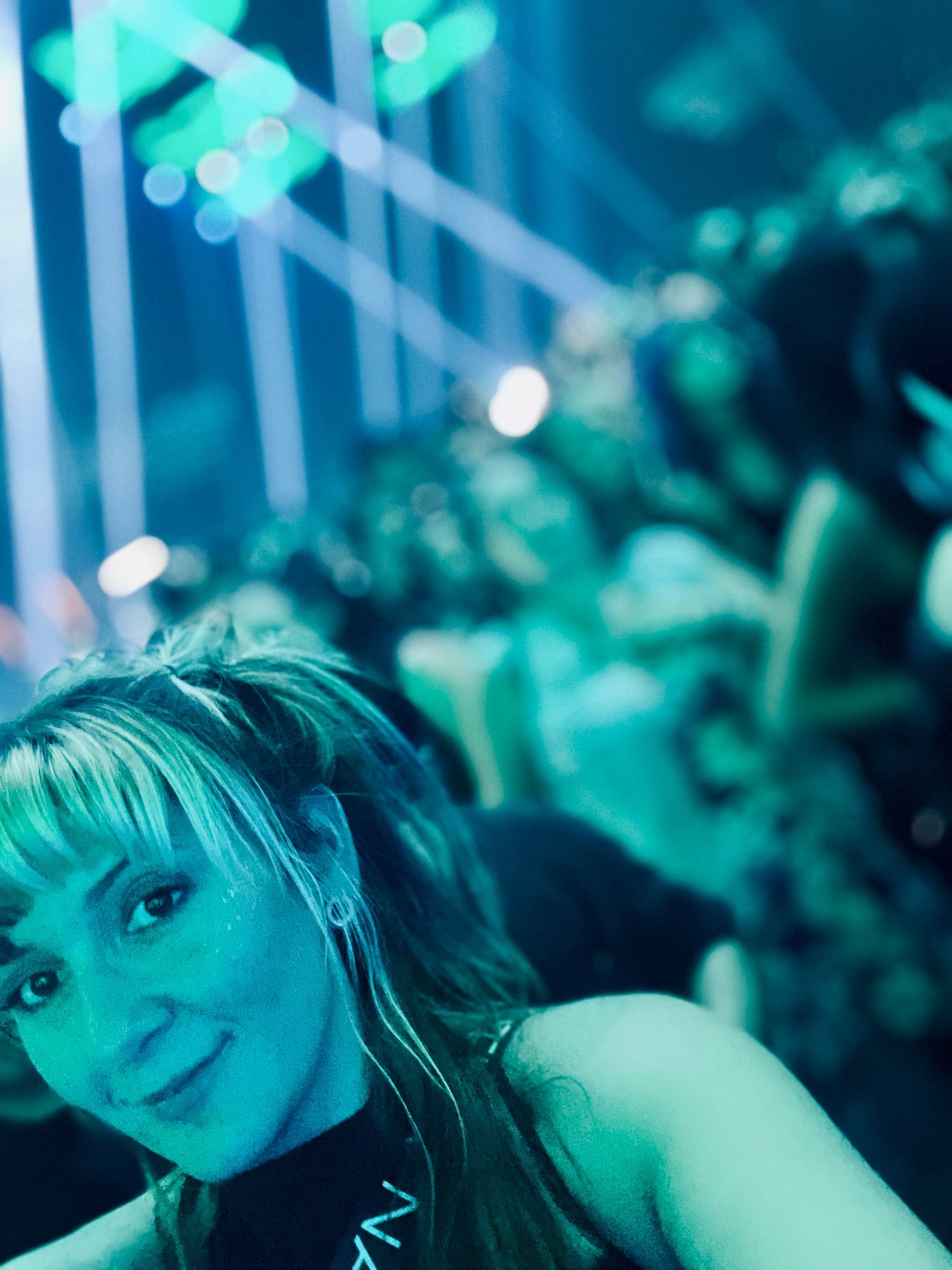 Woman with blonde hair smiling at a concert venue with teal lighting and blurred crowd in background.
