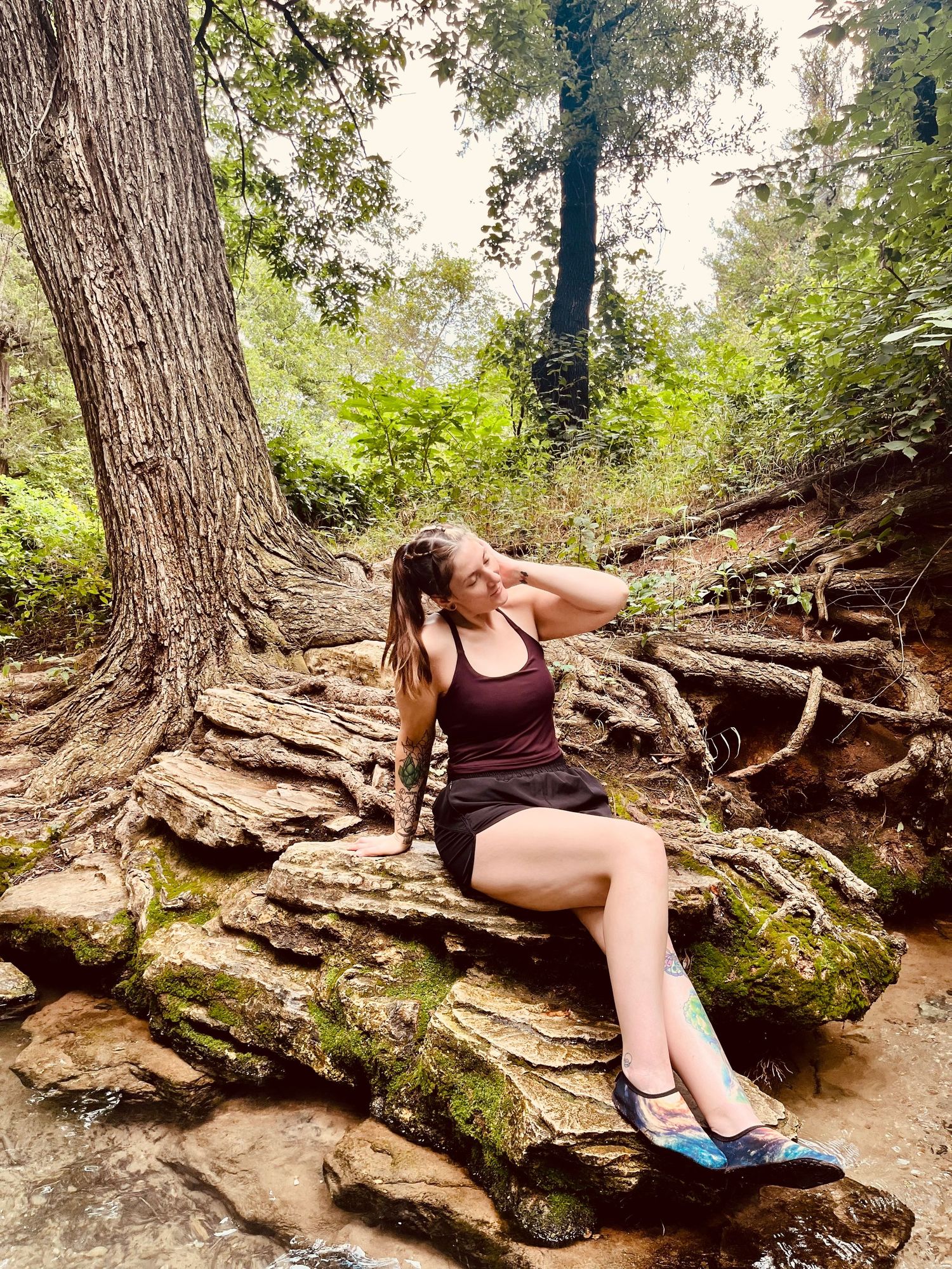 Woman in dark tank top and shorts sits on rocky forest floor beside large tree roots, wearing blue water shoes.