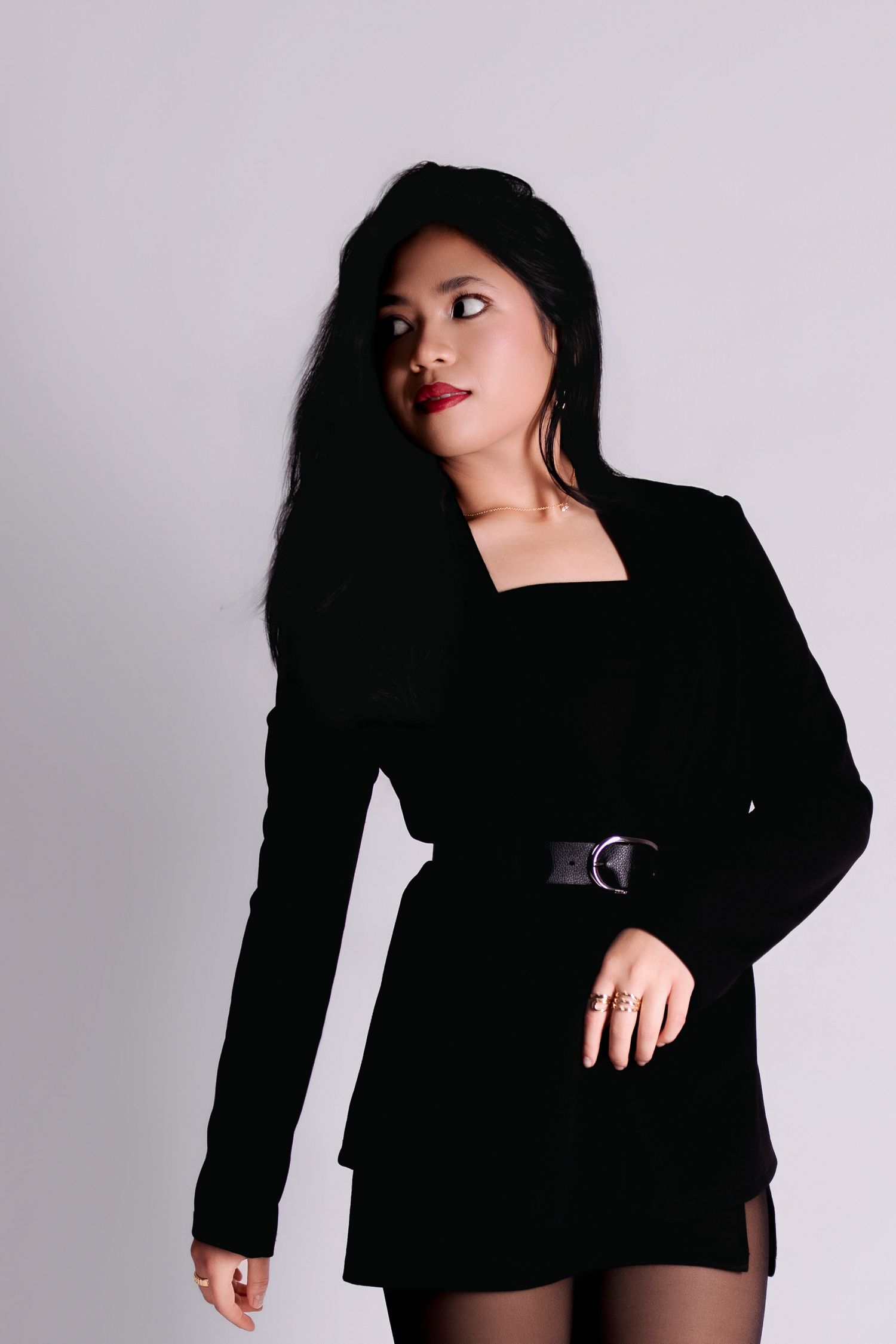 Woman in black belted blazer dress with red lips and long dark hair posing against a light gray background.