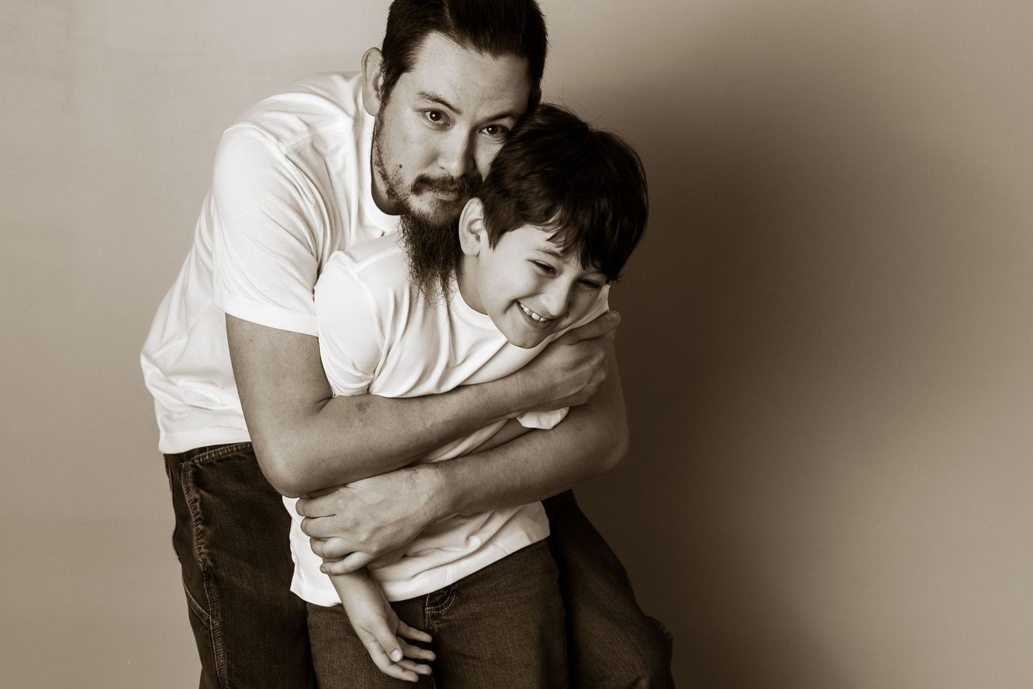 A bearded man in a white t-shirt hugs a young boy from behind in a warm sepia-toned portrait photo.