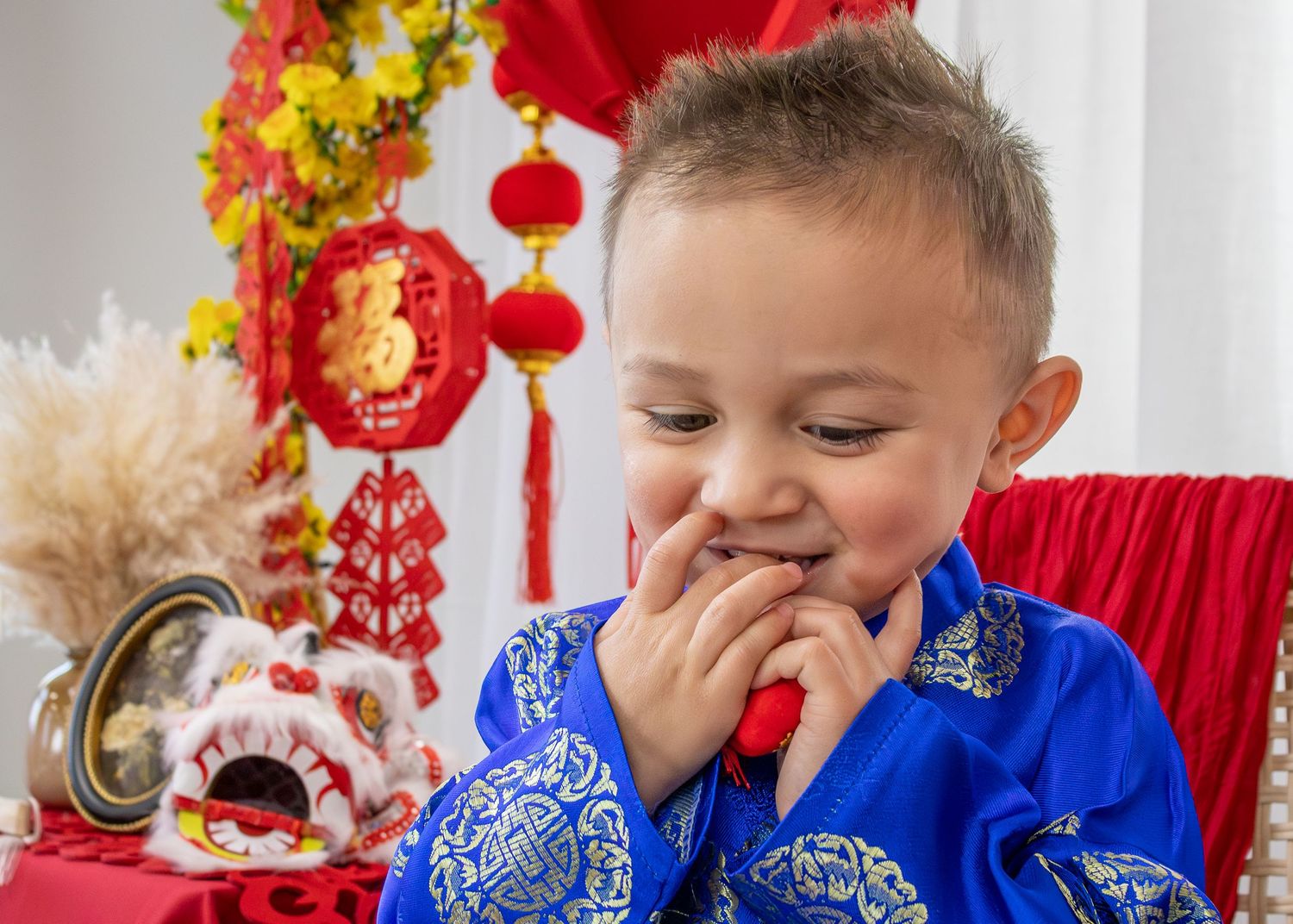 Young boy in blue Chinese traditional outfit smiling during Lunar New Year celebration with red decorations.