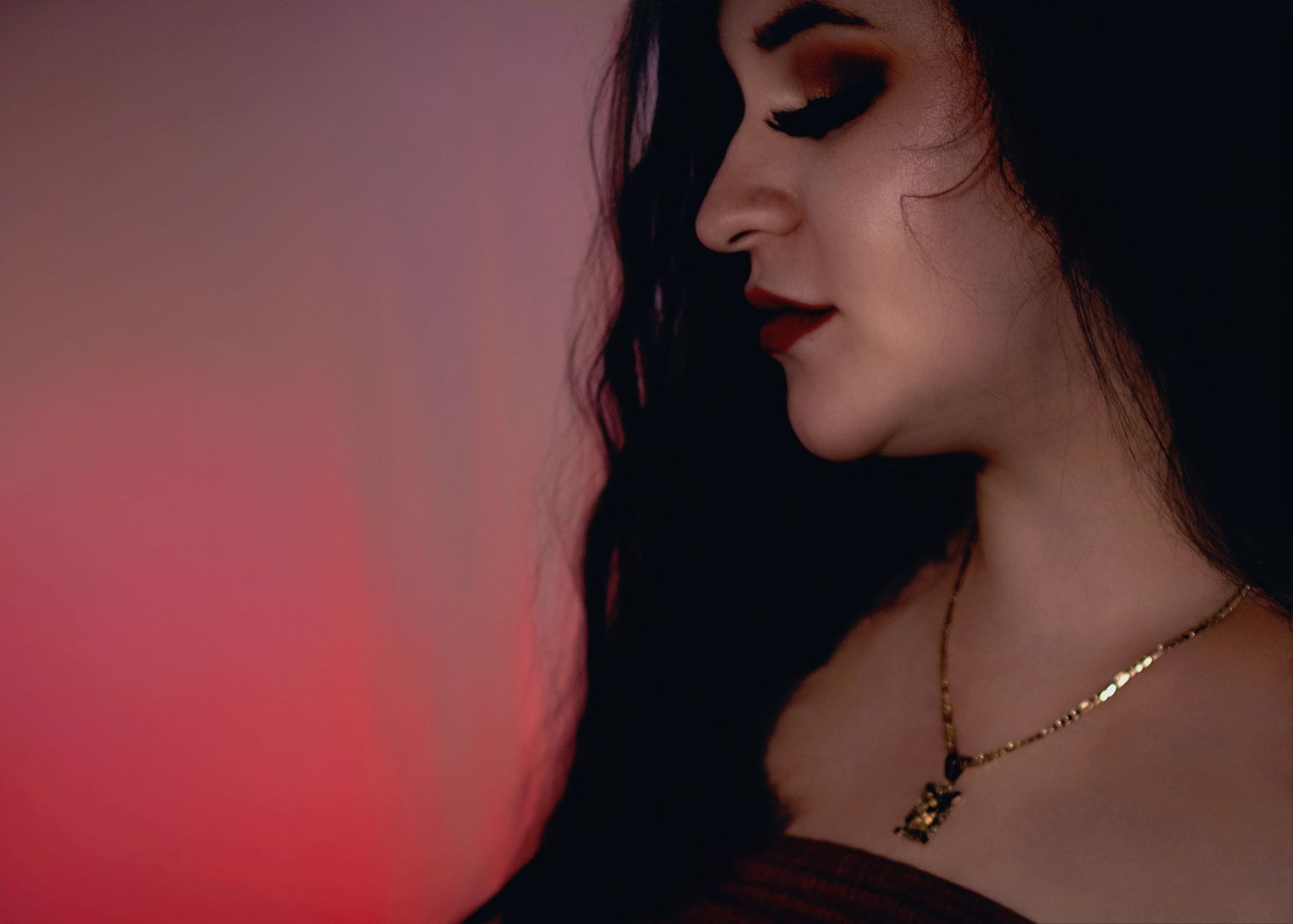 A woman in profile with dark wavy hair, bold red lips, smoky eye makeup, and a gold necklace against a red backdrop.