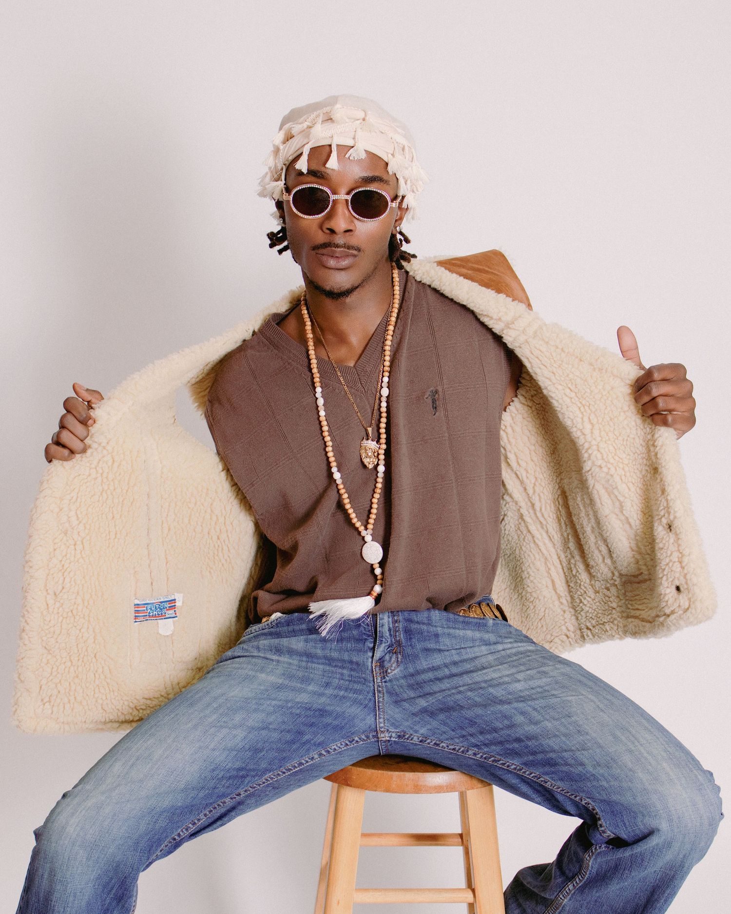 Young man in sherpa jacket, round sunglasses, beaded necklace, and white hat sits on stool giving thumbs up.