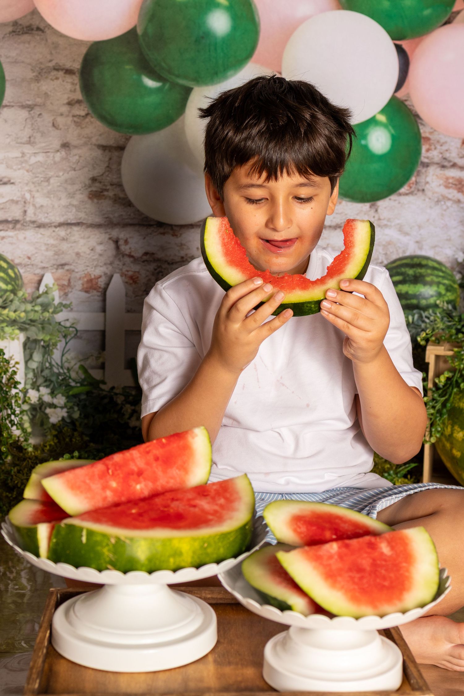A happy boy enjoys eating a fresh watermelon slice at a festive party with green and white balloon decorations.