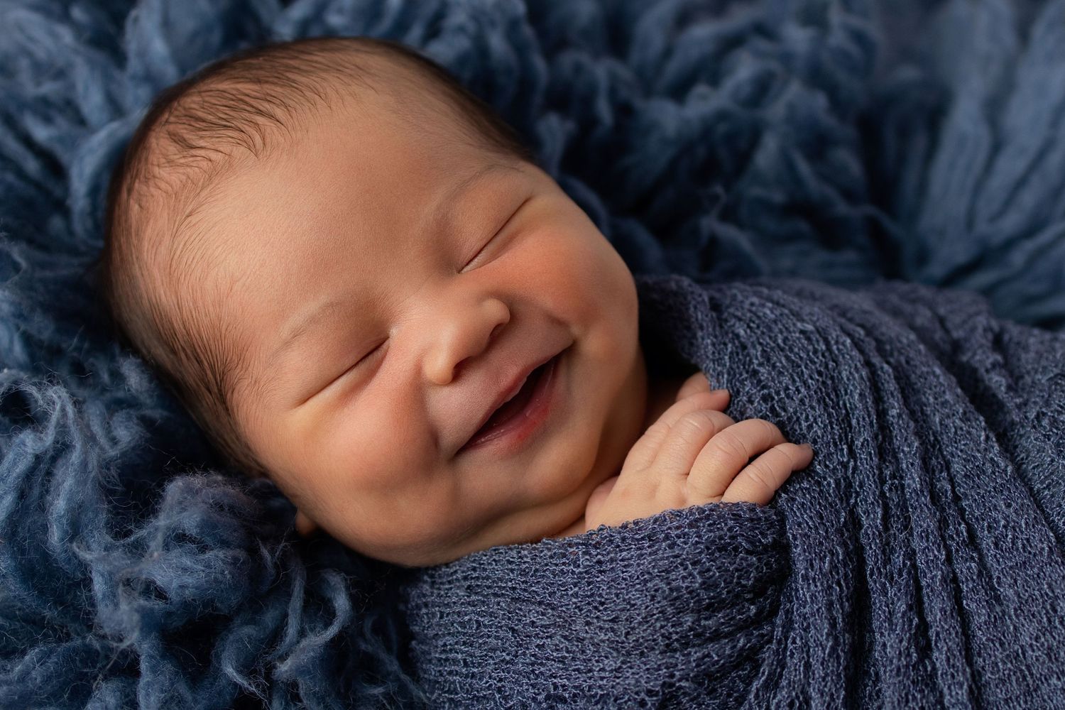 Smiling newborn baby wrapped in blue knit swaddle blanket, sleeping peacefully on soft blue fabric.