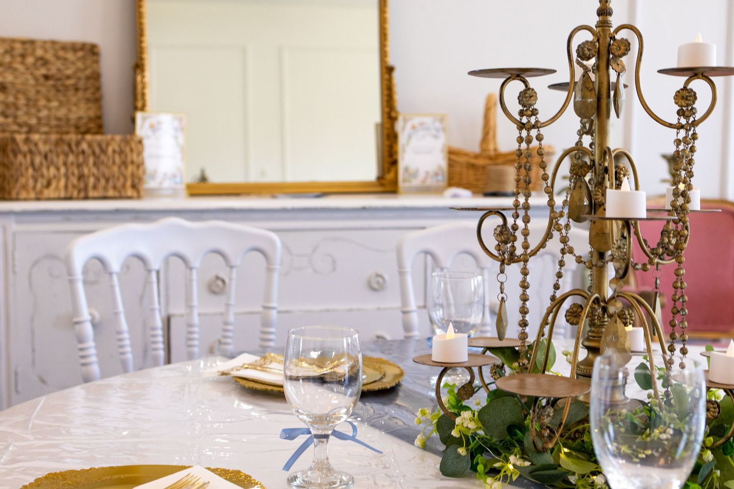 Elegant dining table with candelabra centerpiece, wine glasses, gold accents, and greenery near a white vintage sideboard.