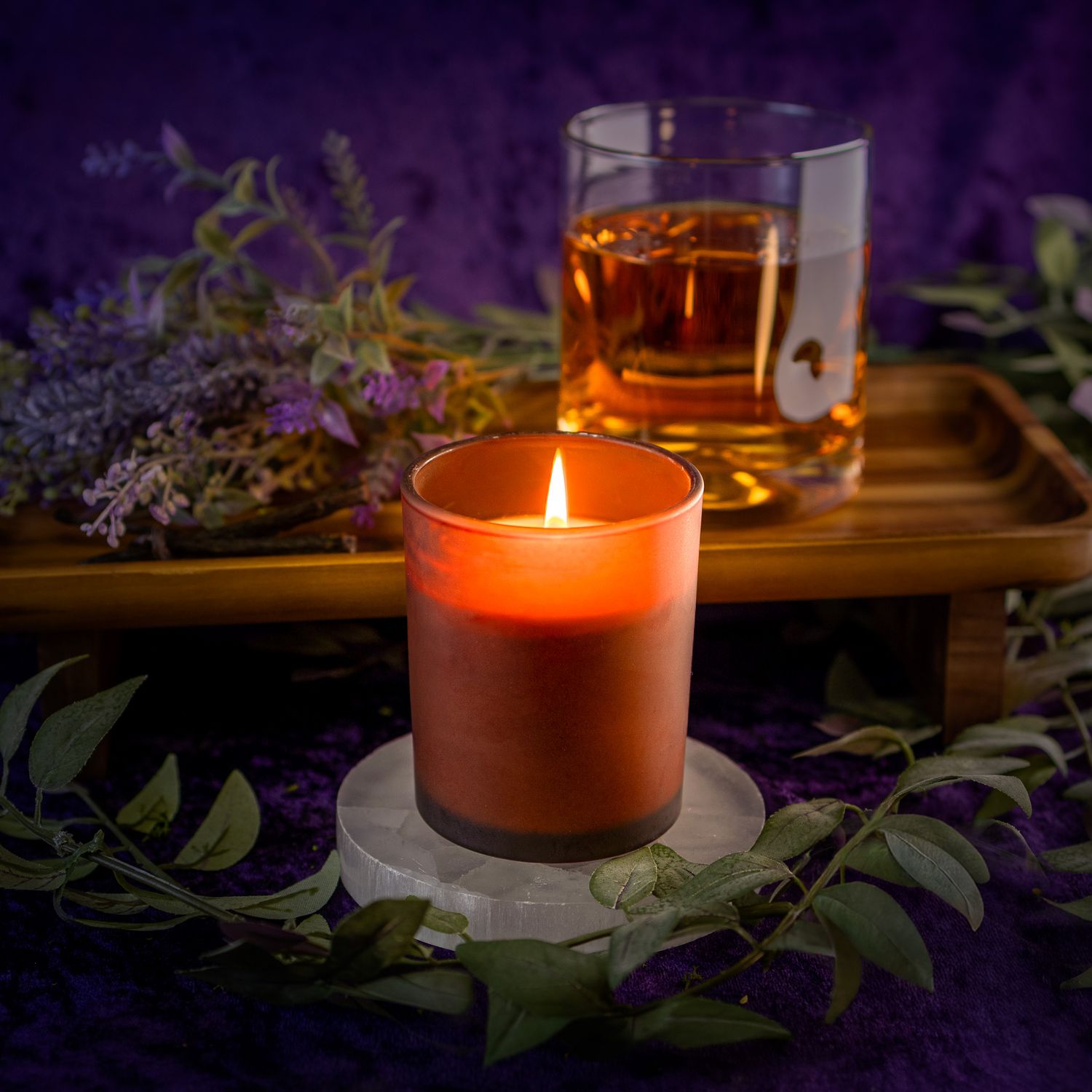 Glowing amber candle surrounded by purple flowers and greenery in dark setting.