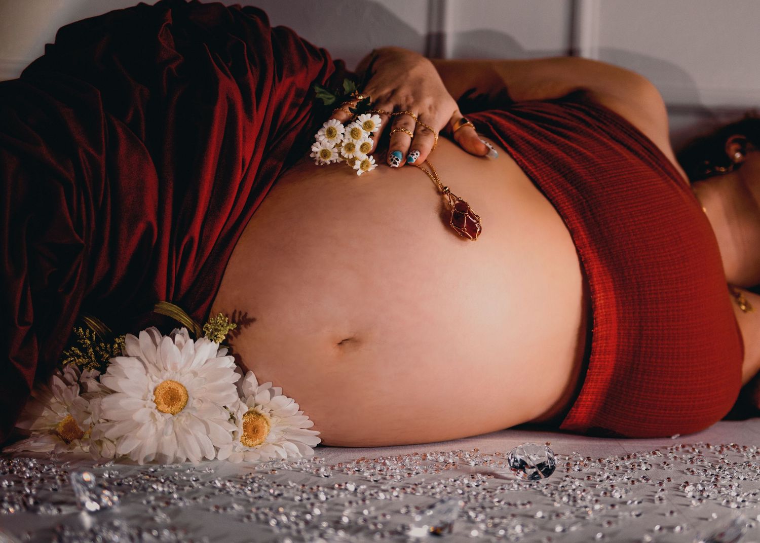 A maternity photoshoot featuring a pregnant belly adorned with white daisies and pearls against a burgundy backdrop.