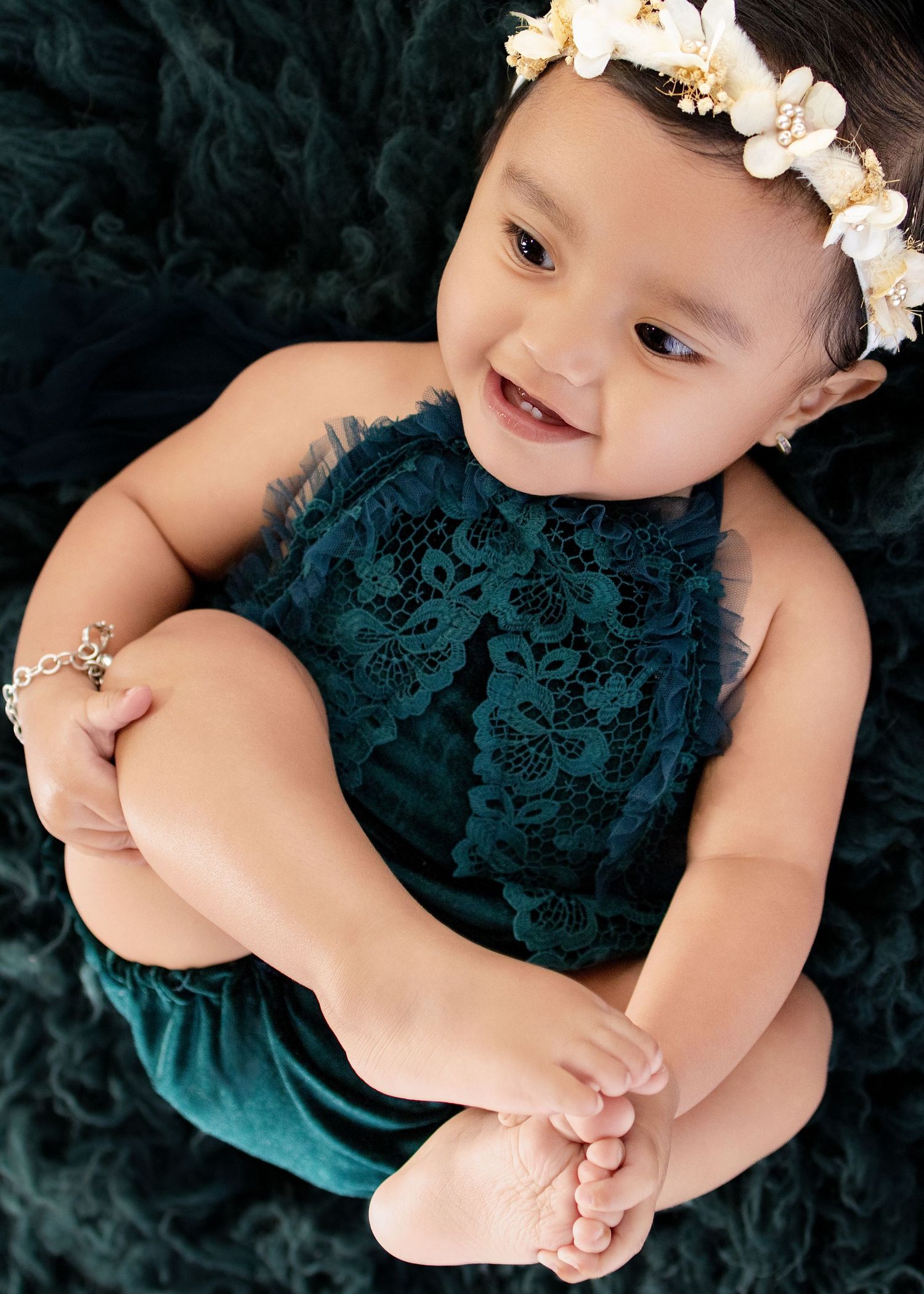 Smiling baby girl in teal lace romper and floral crown sits on dark green fluffy blanket.