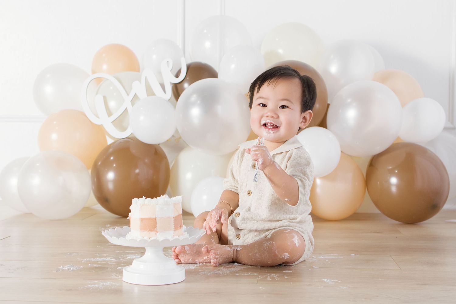 A smiling baby celebrates their first birthday with a white cake and neutral-toned balloons in a bright studio setting.
