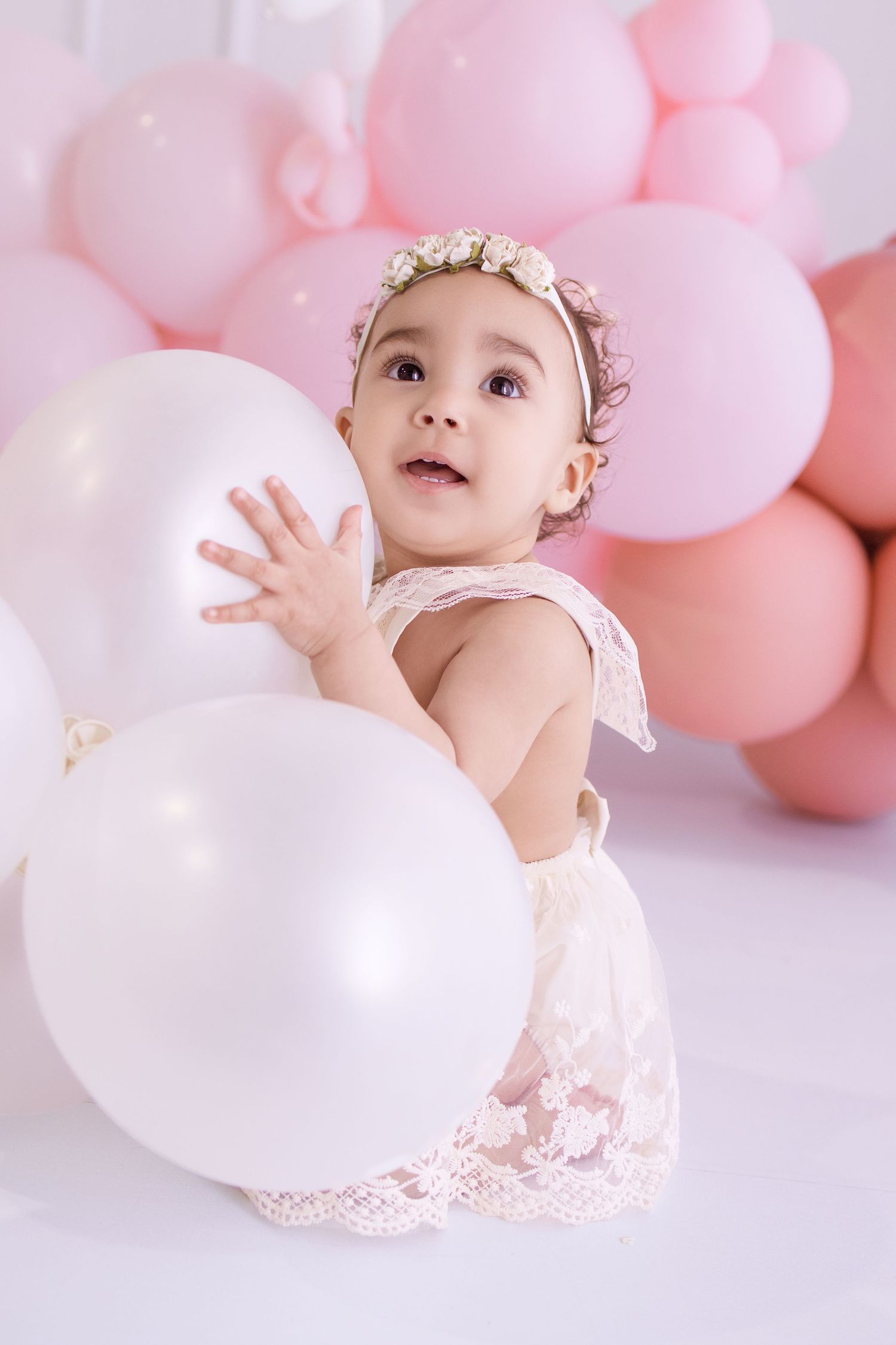 A baby girl in a white lace dress and floral headband plays with white balloons surrounded by pink balloon decorations.