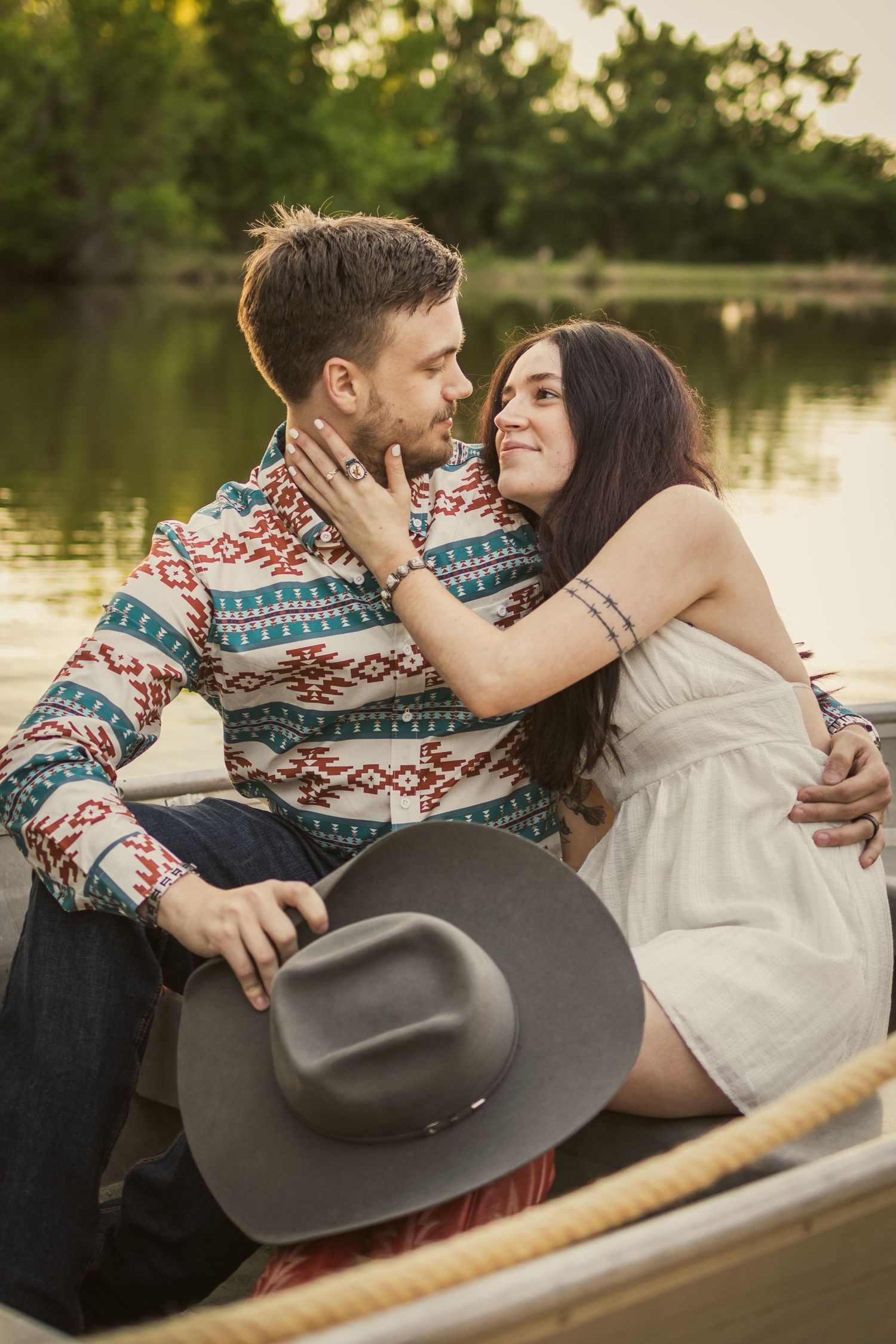 Romantic couple sharing a tender moment in a rowboat by a lake, man in aztec shirt holding cowboy hat.