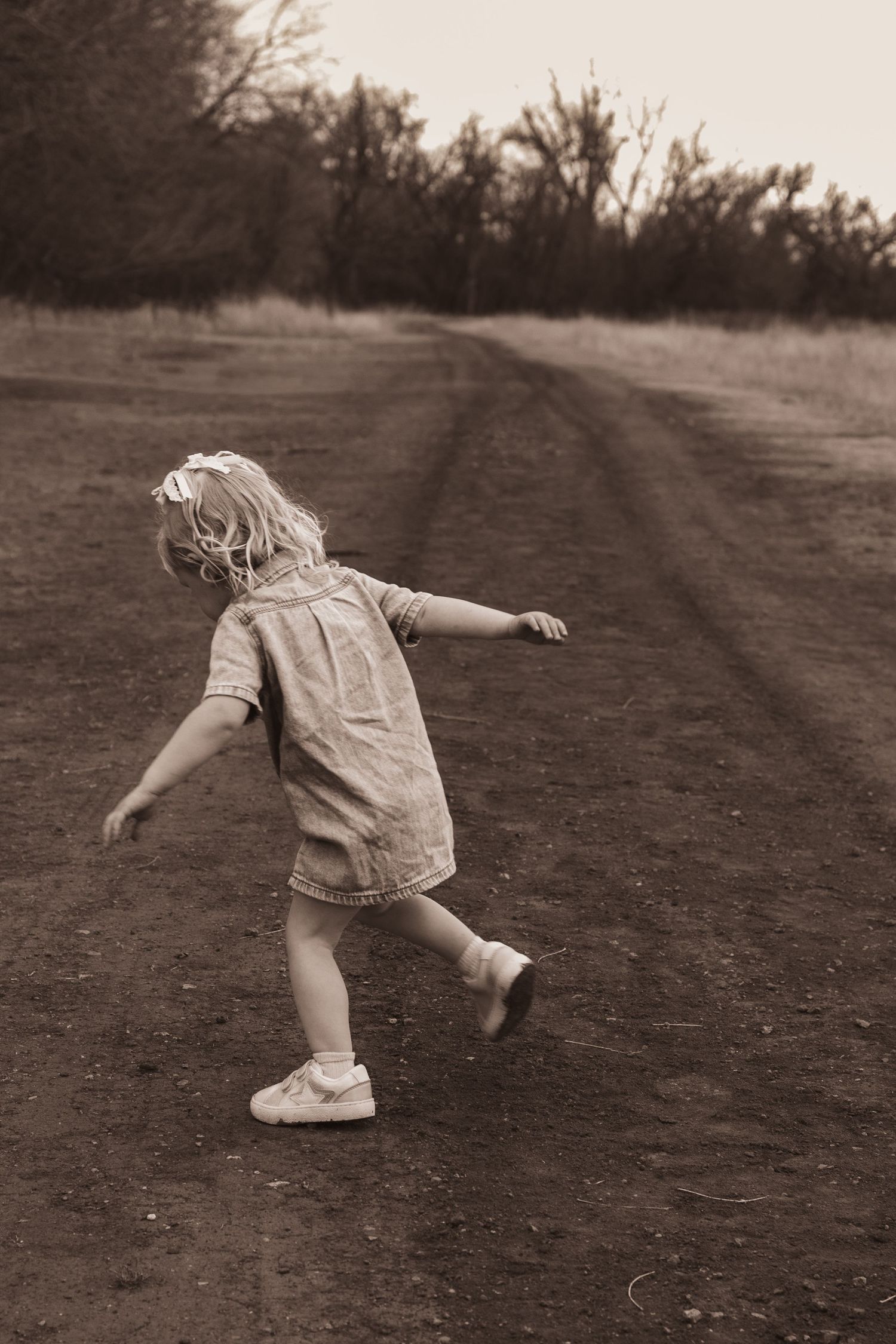 Young girl in denim dress dancing on rural dirt road in sepia toned photo.