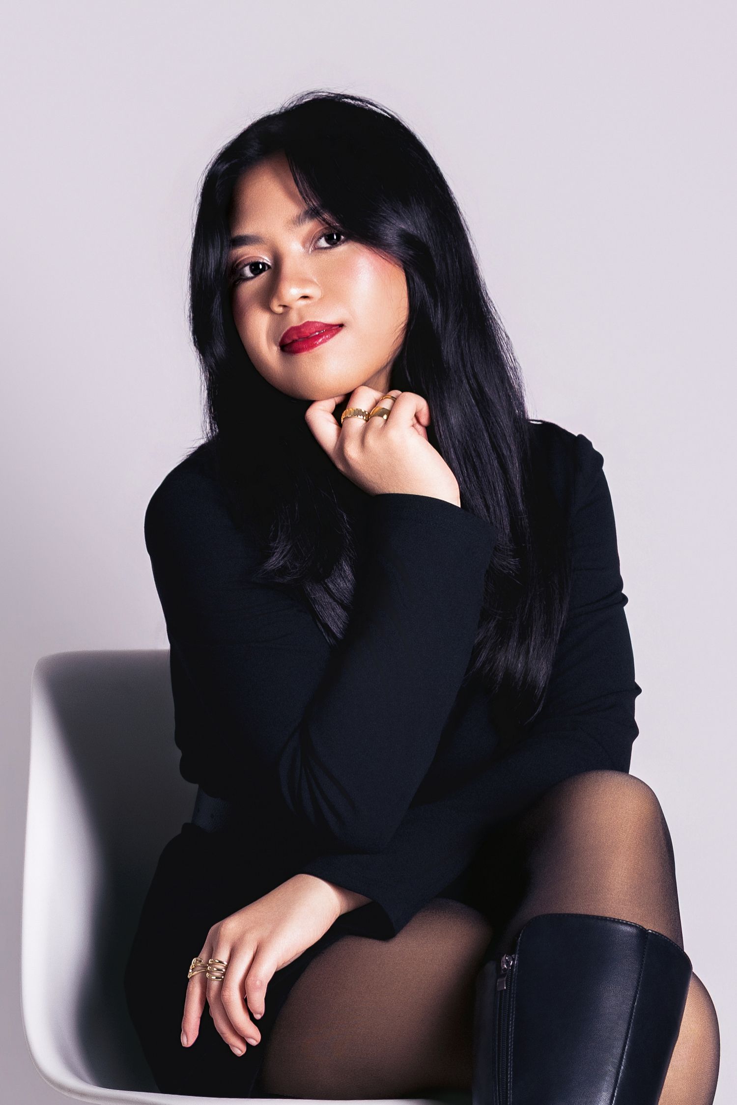 Young Asian woman in black turtleneck and boots sitting elegantly on white chair with red lips.