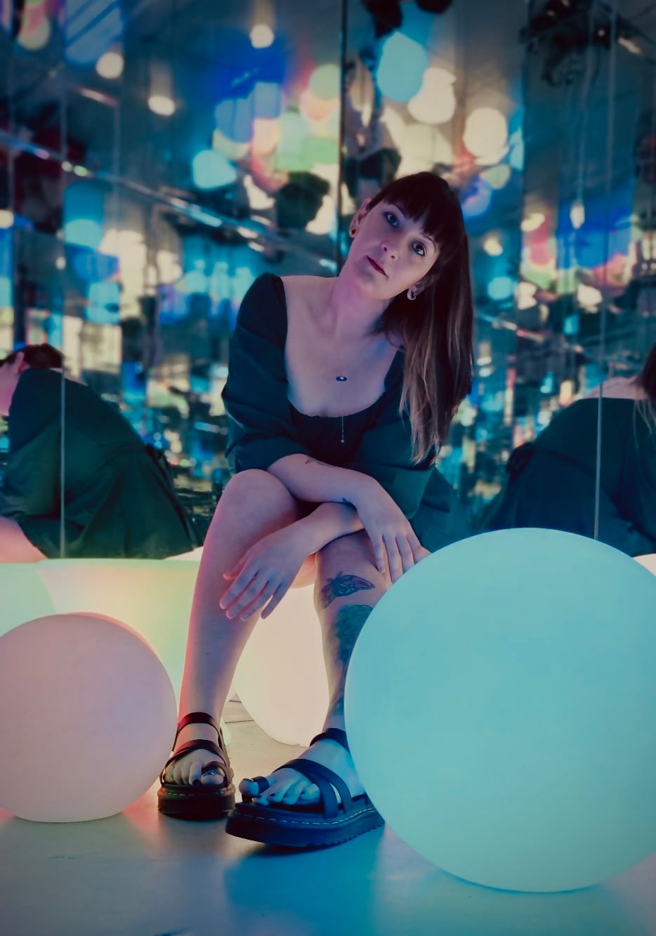 Woman sitting among glowing white orbs in a colorful mirror room with bokeh lights.