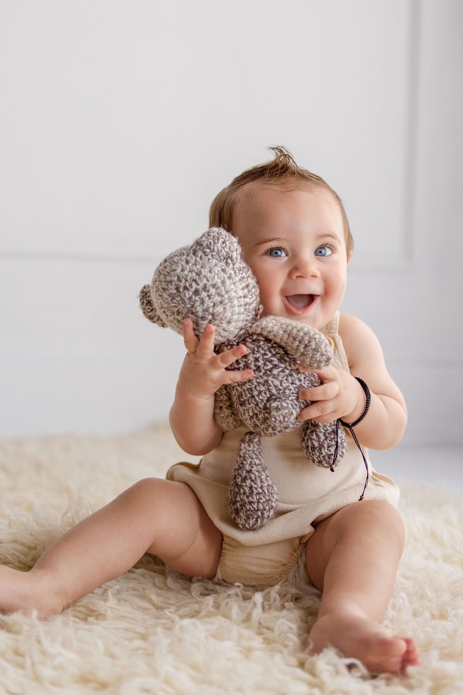 Happy baby in diaper holding crocheted teddy bear while sitting on fluffy cream rug.