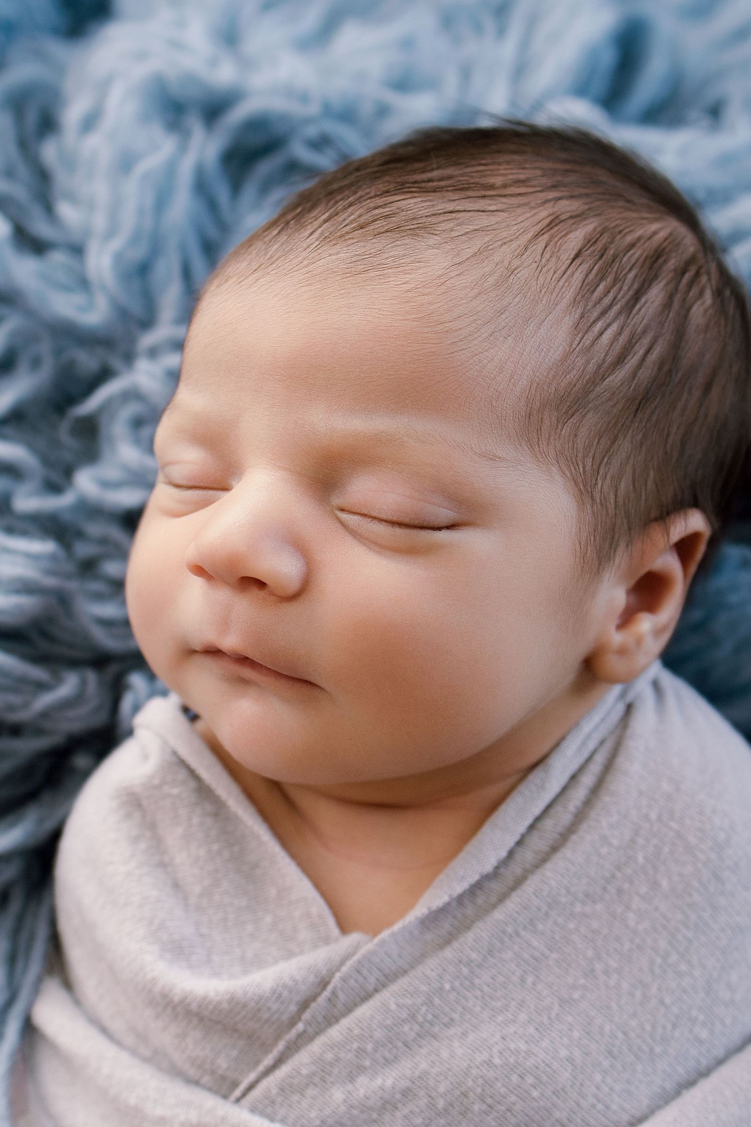 Sleeping newborn baby swaddled in gray wrap lying on soft blue fluffy blanket.