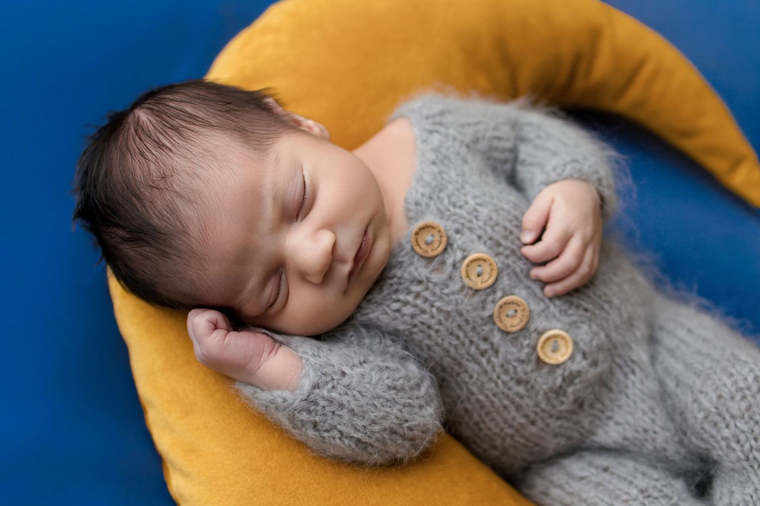 Sleeping newborn baby in gray knit romper with buttons, resting on yellow pillow against blue background.