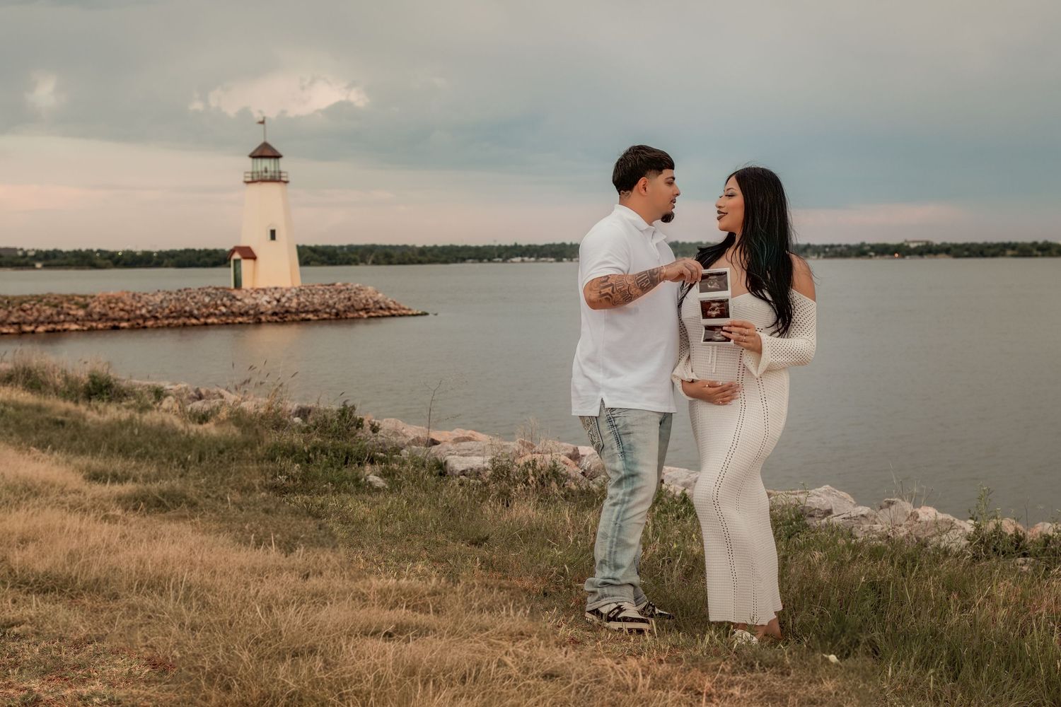 Couple sharing ultrasound photos near a lighthouse by the lake during a maternity photoshoot at sunset.