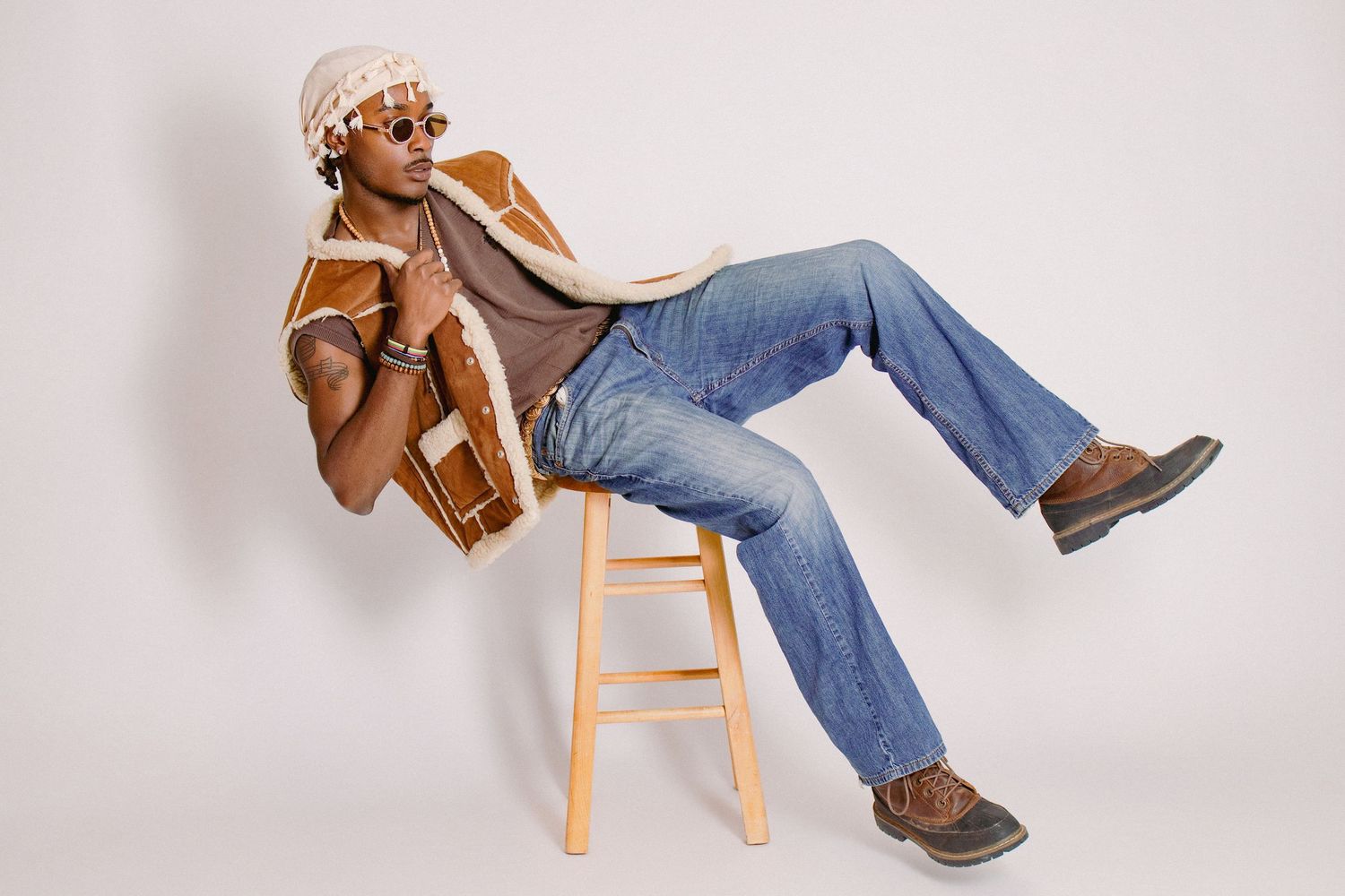 Stylish Black man in shearling vest and flared jeans sits casually on wooden stool wearing sunglasses.