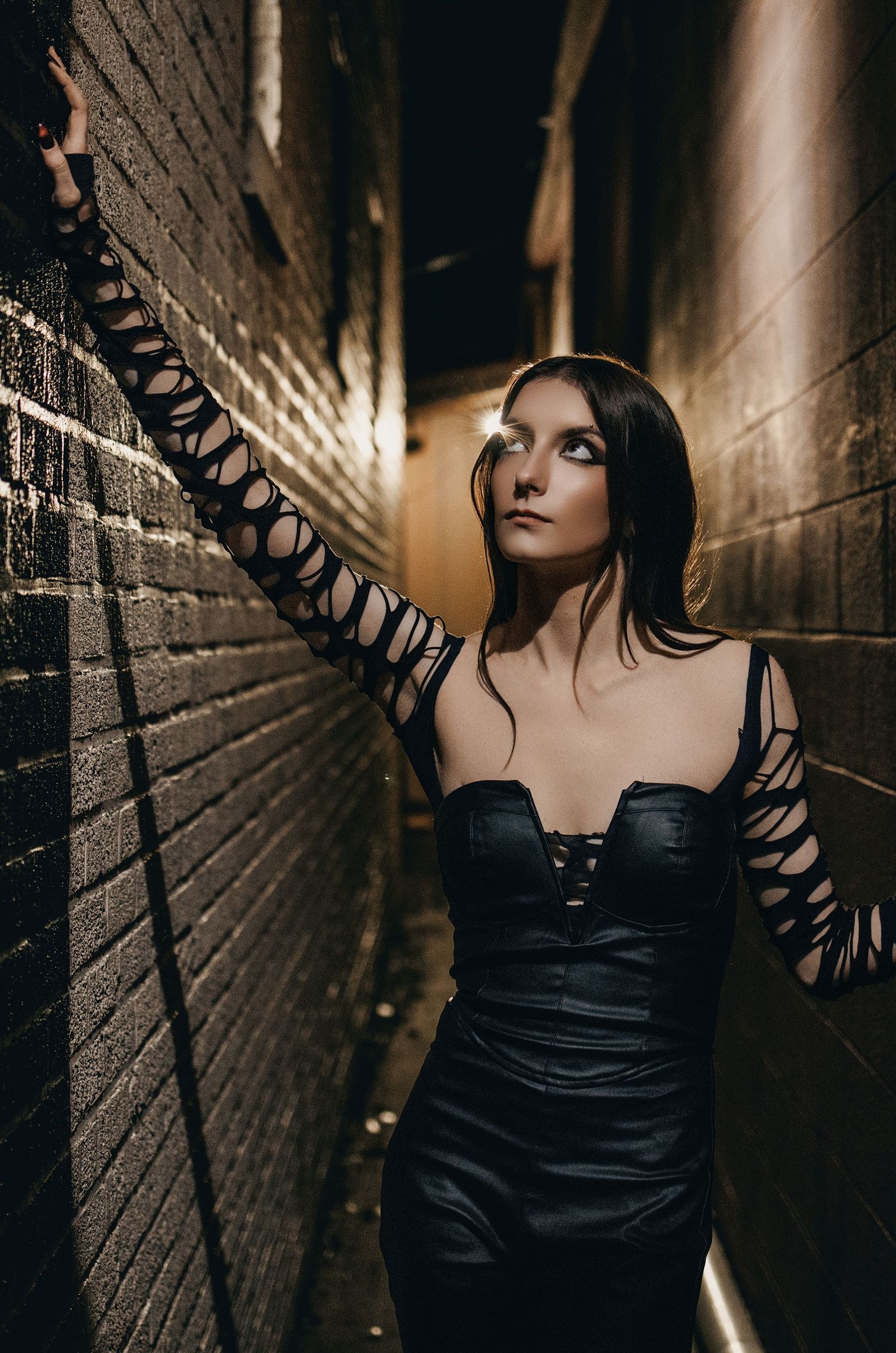 Woman in black corset dress with cutout sleeves posing in brick alleyway.