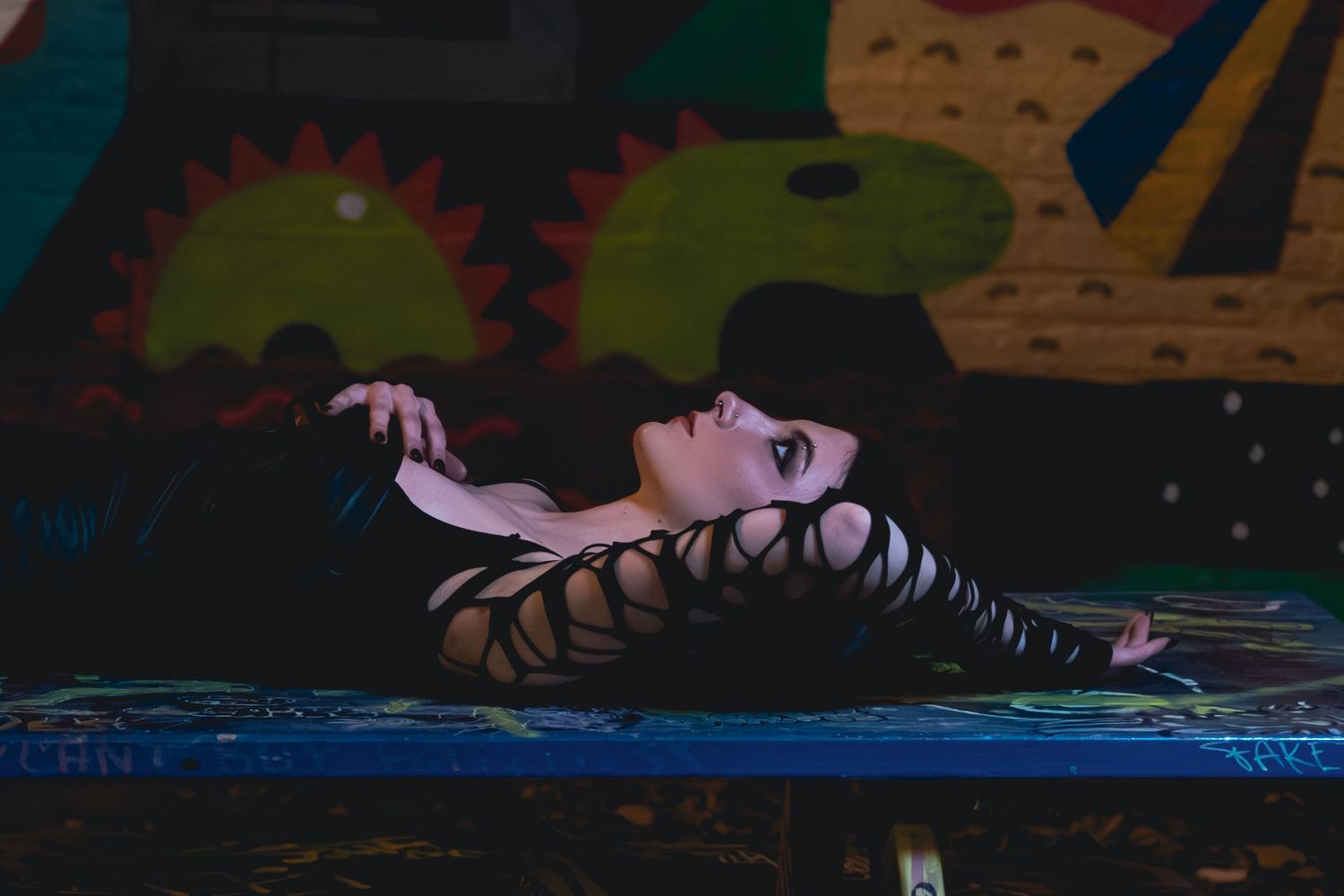 Woman in strappy black outfit lying on a surface against a colorful mural backdrop in dramatic lighting.