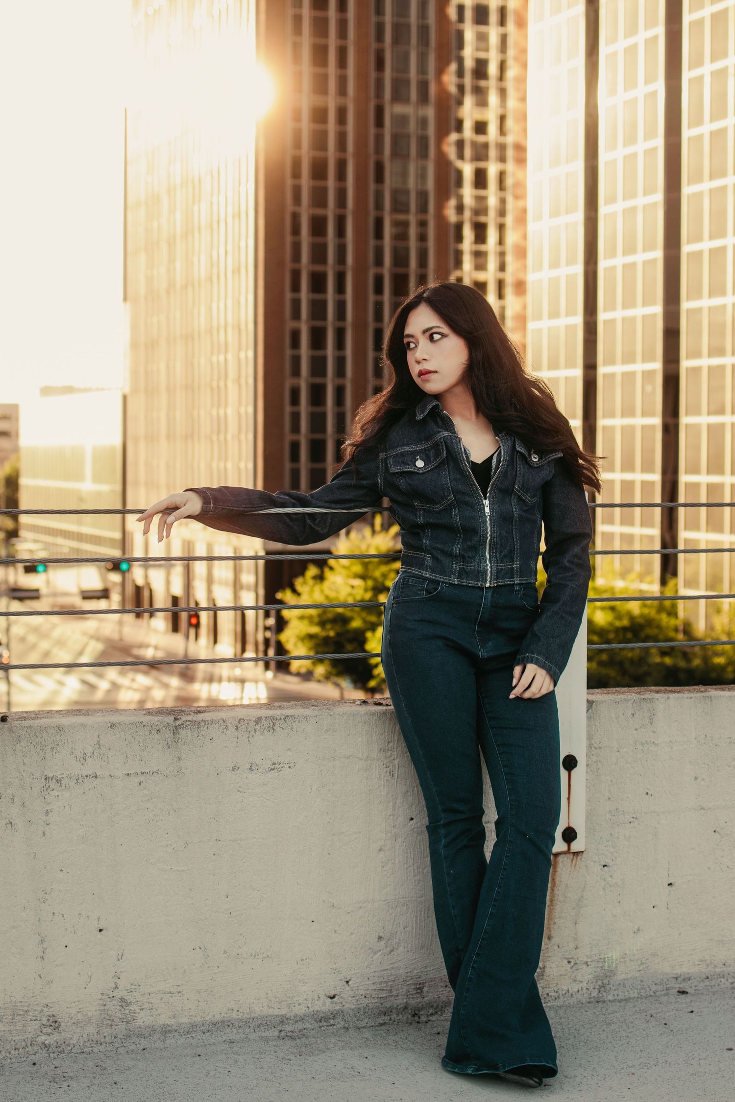 Young woman in dark denim jacket and flare jeans poses on urban rooftop at golden hour with city buildings behind her.