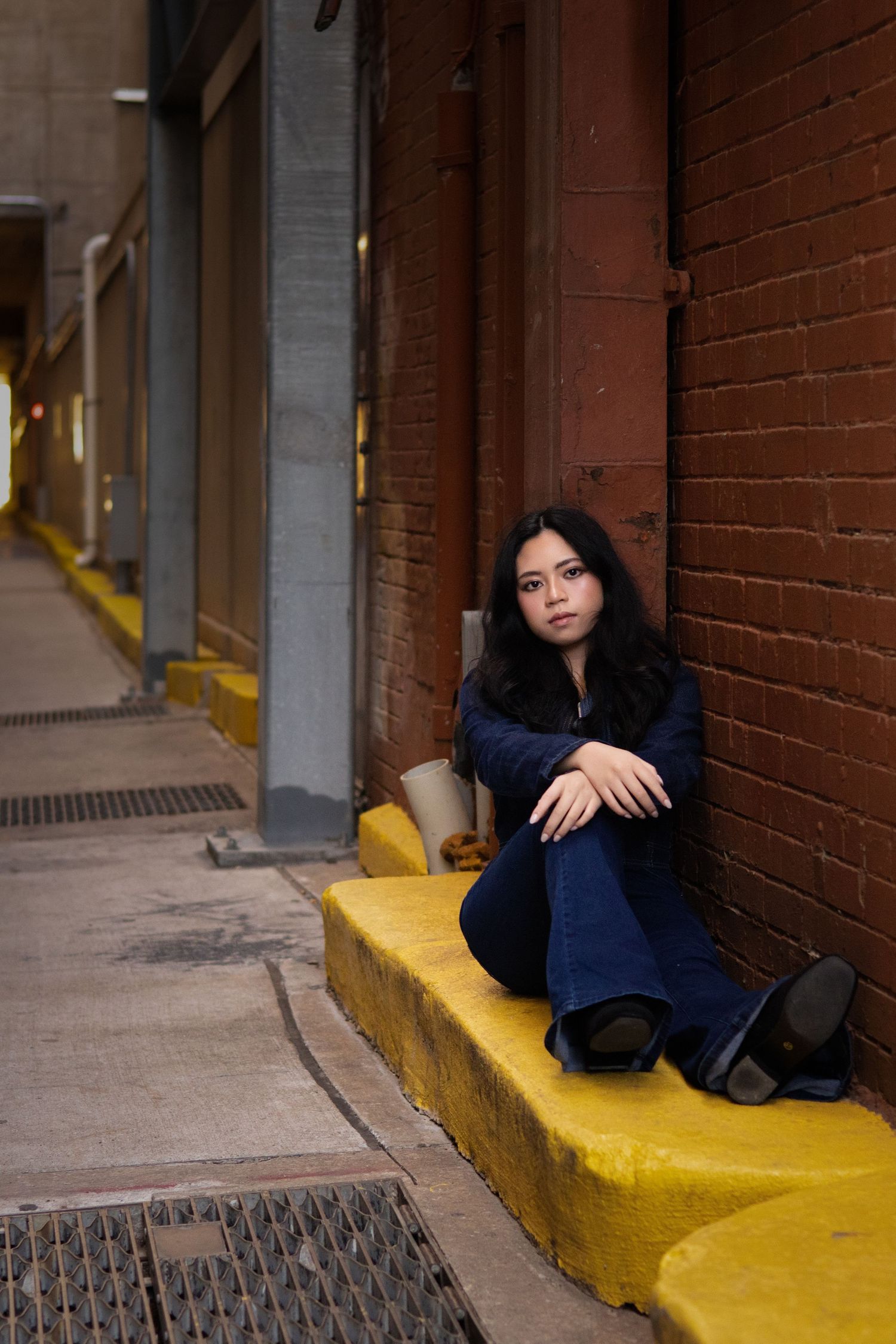 Young woman in navy blue outfit sits on yellow curb leaning against red brick wall in urban alley.