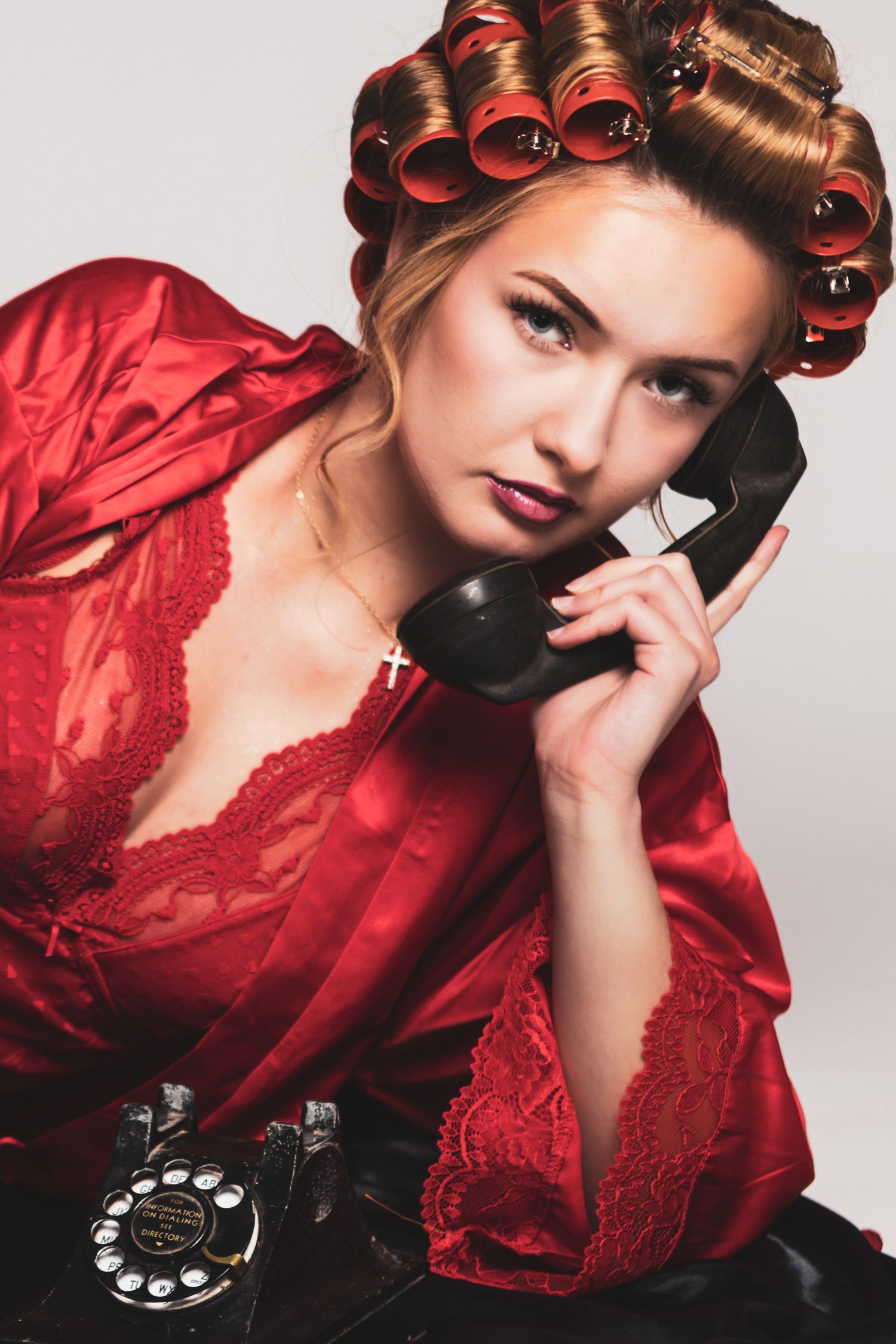 Woman in red satin robe with hair rollers talking on a vintage black rotary telephone.